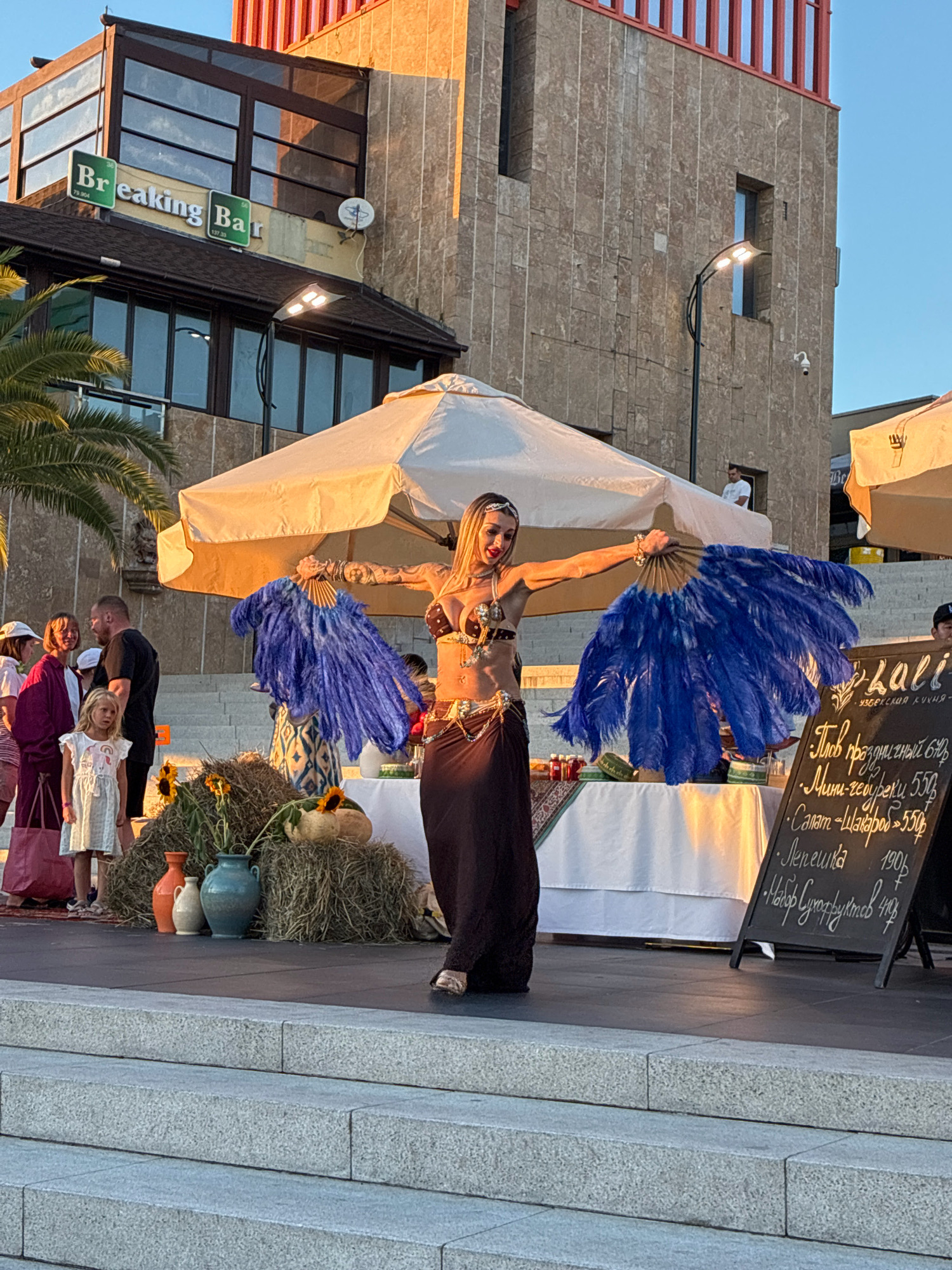 A female belly dancer performs with large blue feather fans in front of a building with a sign that reads "Breaking Bar" in Sochi, Russia. The dancer wears a bejeweled brown top and a long dark skirt. She is positioned on a raised platform, likely outdoors given the sunlight and palm tree visible. Behind her, a white tent provides shade for a table laden with food items, including sunflowers and gourds. Spectators, including children and adults, stand to the left of the performer, observing the show. A black chalkboard menu is visible to the right of the dancer, displaying items and prices in a foreign script. The background features a stone building with large windows and a red architectural detail at the top. The scene is bathed in the warm glow of late afternoon or early evening sun.