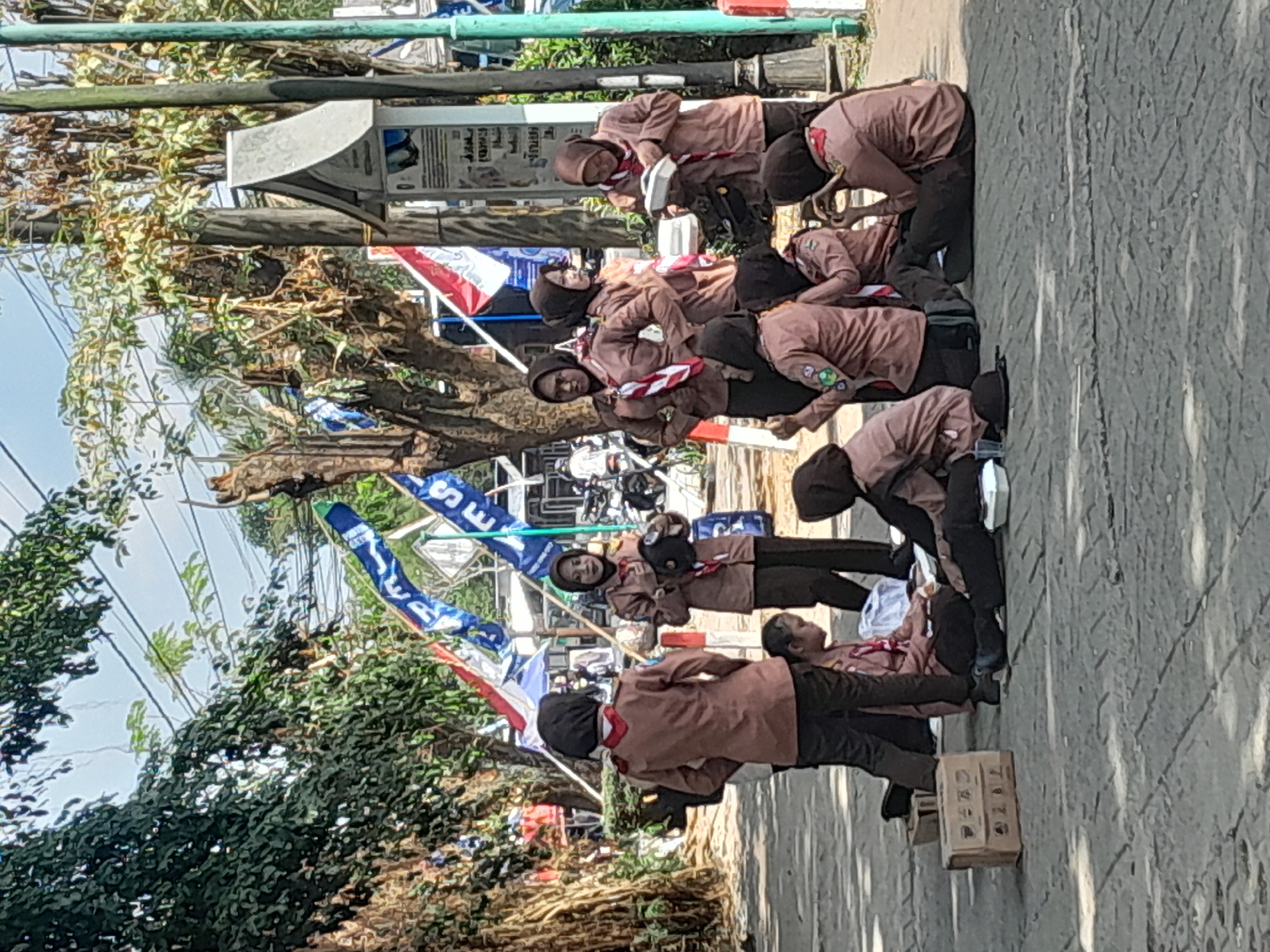 In the city of Genteng Kulon, Indonesia, a group of girls in brown scout uniforms are gathered on a paved area under trees. Some are standing, while others are seated on the ground. Many wear headscarves, some brown and some black. They appear to be participating in an outdoor activity, possibly a scout meeting or event, with several flags visible in the background. A few of the girls are interacting with food containers, suggesting they might be having a meal or snack. The environment includes trees, a sidewalk, and a road with some vehicles in the distance. The overall scene depicts a casual gathering of young people in a public outdoor setting.