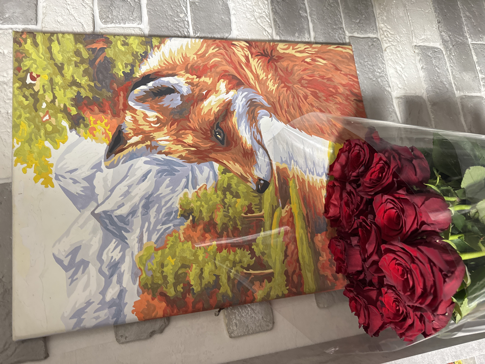 The image shows a painting of a fox with mountains and trees in the background, leaning against a brick wall. In the foreground, a bouquet of red roses is wrapped in clear plastic. The painting is positioned vertically, and the roses are arranged in front of it, partially obscuring the bottom of the painting. The scene appears to be indoors, possibly in a home or a gift shop, given the presence of both decorative items. The brick wall is painted in shades of gray and white, suggesting a textured surface. There is no indication of the location being in Chaikovsky, Russia.