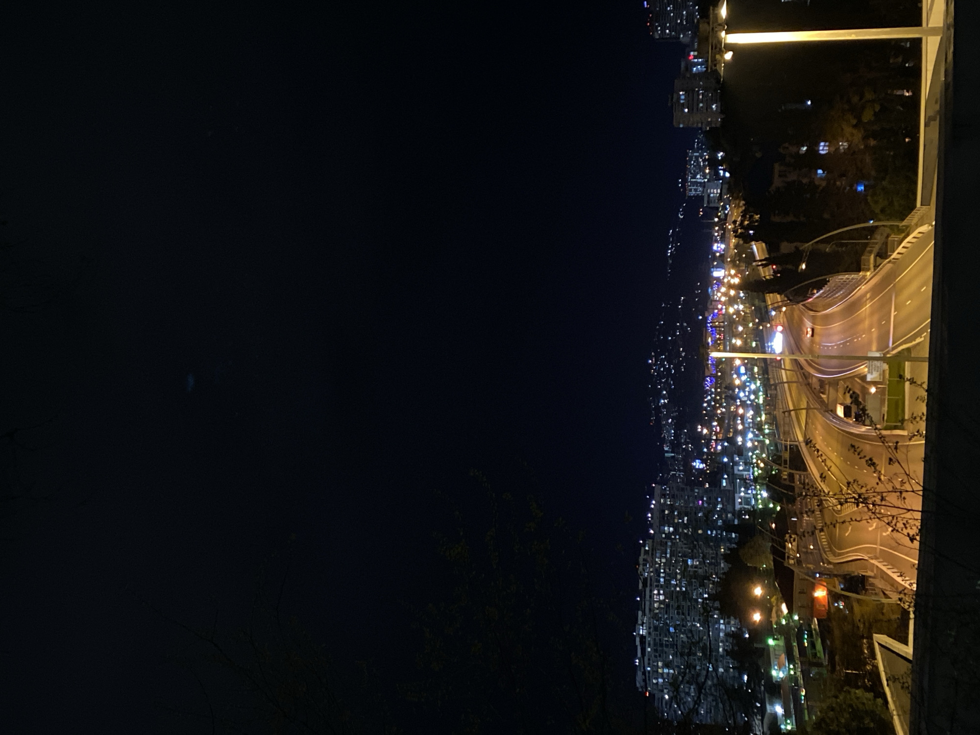 This media file presents a night view of a city, identified as Sochi, Russia, captured from an elevated perspective. The scene is dominated by a sweeping urban landscape illuminated by countless artificial lights against a dark, featureless sky.
In the mid-ground, a multi-lane highway or major road curves gracefully from the right foreground towards the center-left, stretching into the distance. It is brightly lit by numerous streetlights, casting a warm glow on the asphalt and highlighting the distinct white and yellow lane markings. A solitary red vehicle is faintly visible on the road, indicating ongoing, albeit light, traffic.
Beyond the road, the cityscape unfolds, a dense array of buildings whose windows glow with a multitude of lights—predominantly yellow, white, and blue—suggesting active residential and commercial areas. These lights create a vibrant, mosaic-like pattern that extends far into the background, hinting at the vastness of the urban development. Silhouetted trees and sparse vegetation frame the bottom edges of the image, barely discernible against the bright city lights.
The time of day is clearly night, as evidenced by the deep black sky and the reliance on artificial illumination. No specific weather conditions are discernible, suggesting a clear night. There are no visible people or specific events taking place, but the illuminated city evokes a sense of constant activity and urban energy. No discernible text is visible within the image.