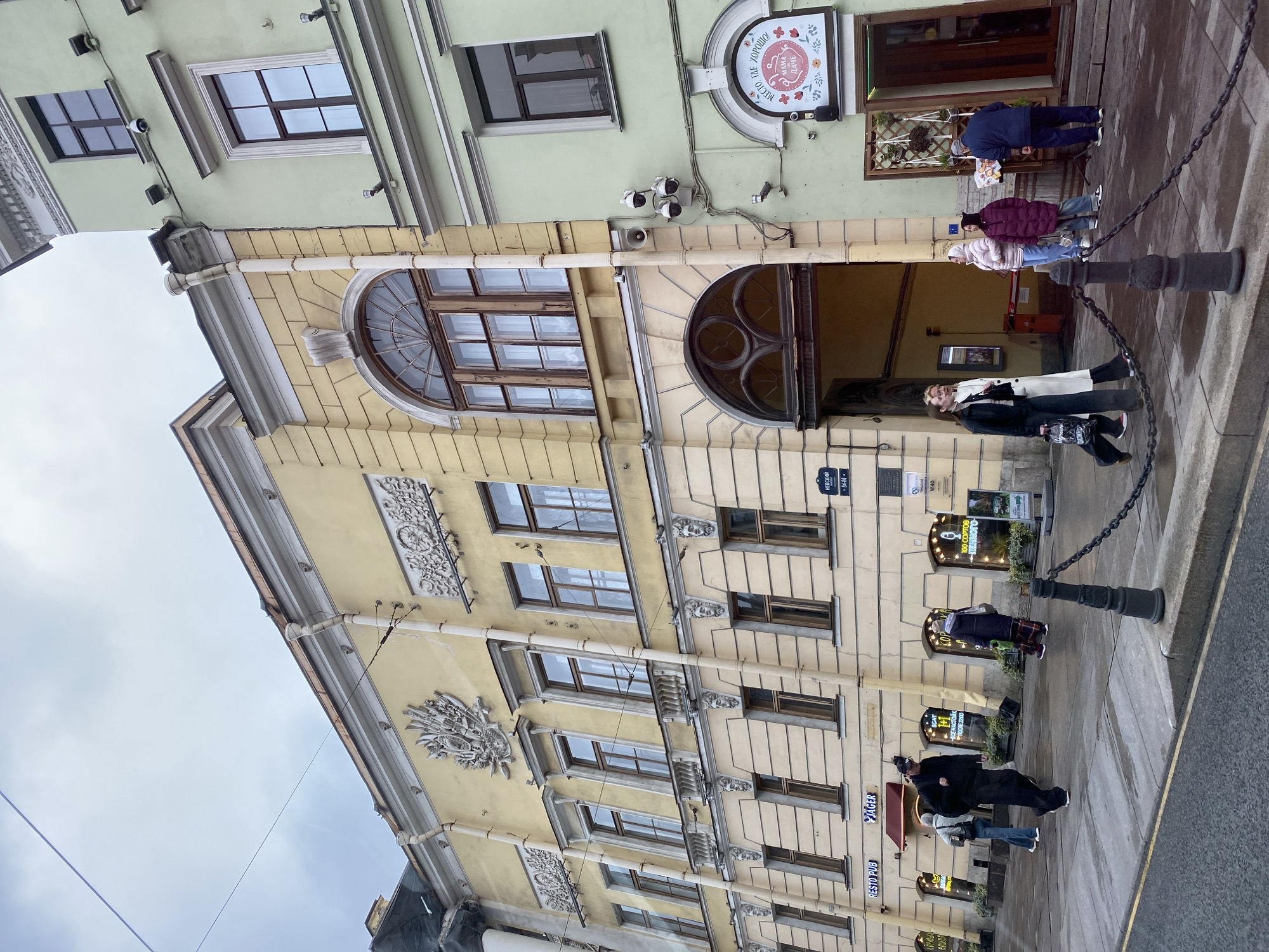 Here is a description of the image:
The image depicts a street scene in an urban environment, likely in Saint Petersburg, Russia, based on the Cyrillic signage. The architecture features buildings with a light yellow facade and decorative elements. The street is wet, suggesting recent rain. People are present, walking and standing near the entrance of what appears to be a pub or restaurant called "JÄGER RESTO PUB." A sign also indicates another establishment called "MAMA ДАЧЕ." Street signs with Cyrillic text are visible, including "НЕВСКИЙ" and a street number. A chain is strung between posts along the sidewalk. The sky appears overcast.