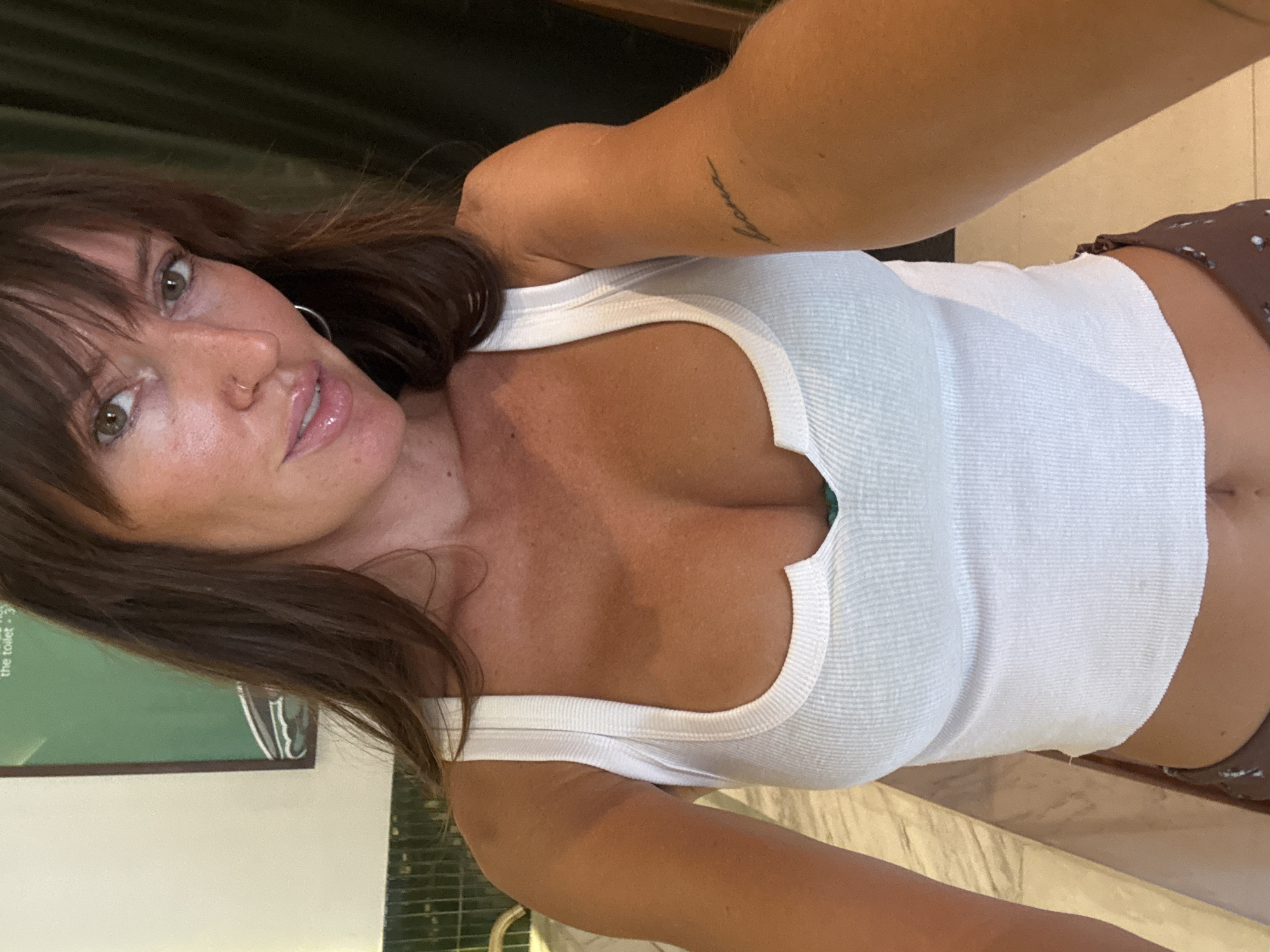 A woman with brown hair and green eyes is taking a selfie in what appears to be an indoor setting. She is wearing a white tank top. Her right arm is raised, holding the camera. She has a nose ring and an earring. The background is somewhat blurred, but it includes a green wall with text on it and a light-colored surface. The location is Mengwi, Indonesia.