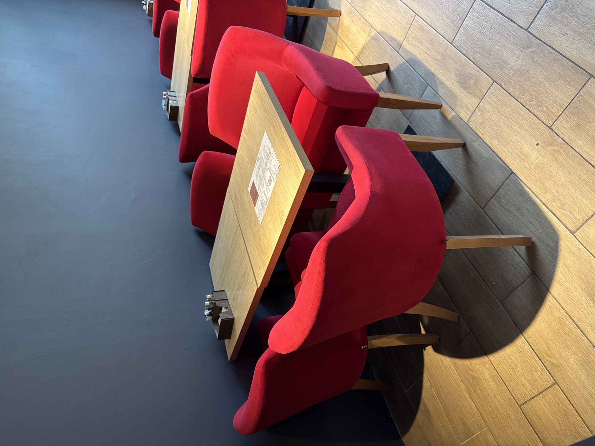 The image shows a row of red armchairs with wooden tables between them. The scene appears to be in a cafe or restaurant. The floor is dark grey on one side and light wood on the other. The image orientation suggests a view looking down the length of the tables and chairs from an elevated perspective. Based on the architectural style, it's likely the photo was taken in Saint Petersburg, Russia. There are no people visible in the frame.
