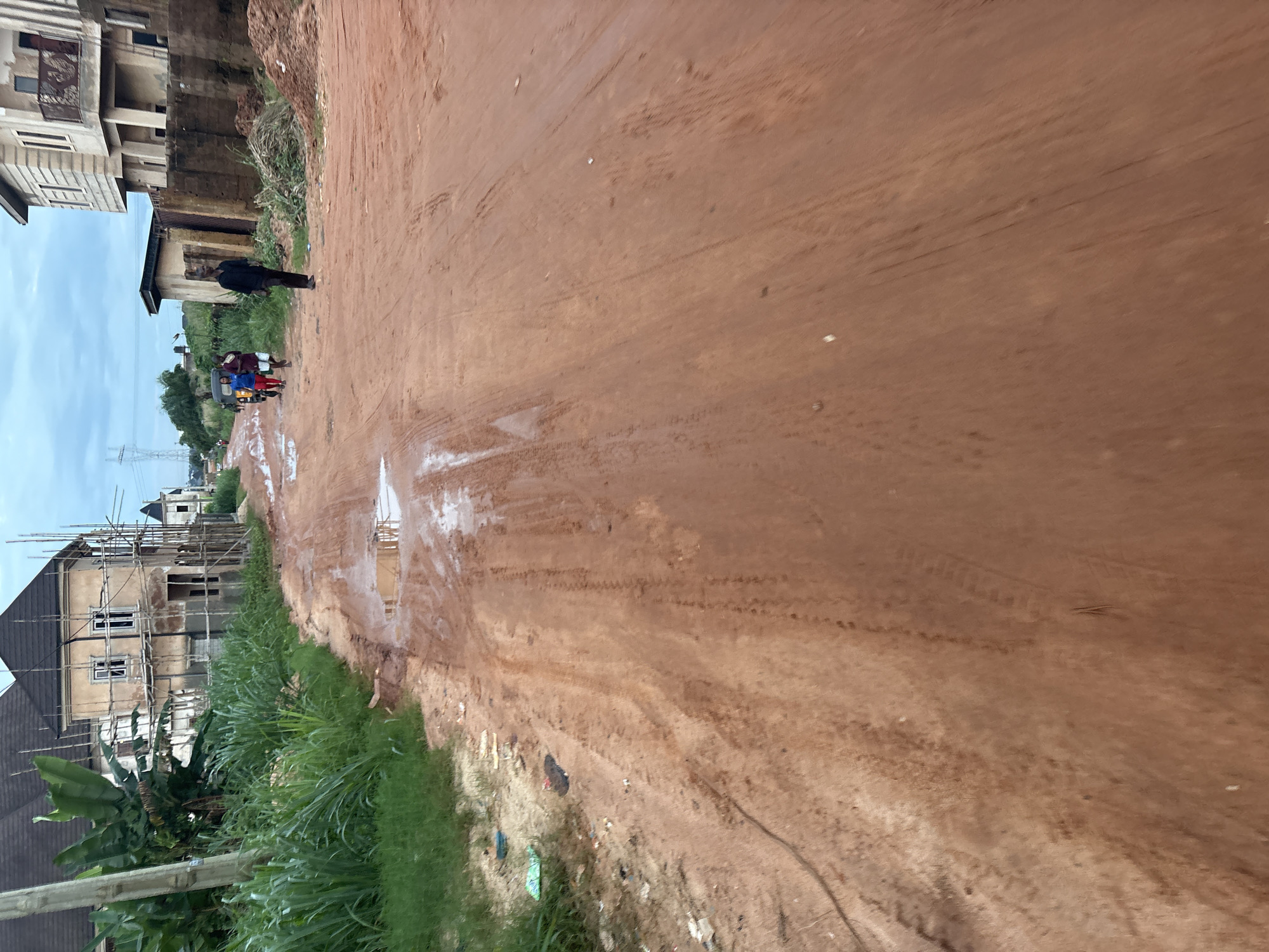 A low-angle, full shot captures a red dirt road in Obosi, Nigeria, under an overcast sky. The road is muddy and wet, with several puddles reflecting the sky. On the left side of the road, there is lush green grass and a wooden pole. In the distance, unfinished houses with scaffolding are visible. Several people are walking on the road, including a man in a black shirt and dark pants, and a group of three individuals further down the road. A motor tricycle is also present. The scene suggests an ongoing construction project in a developing area.