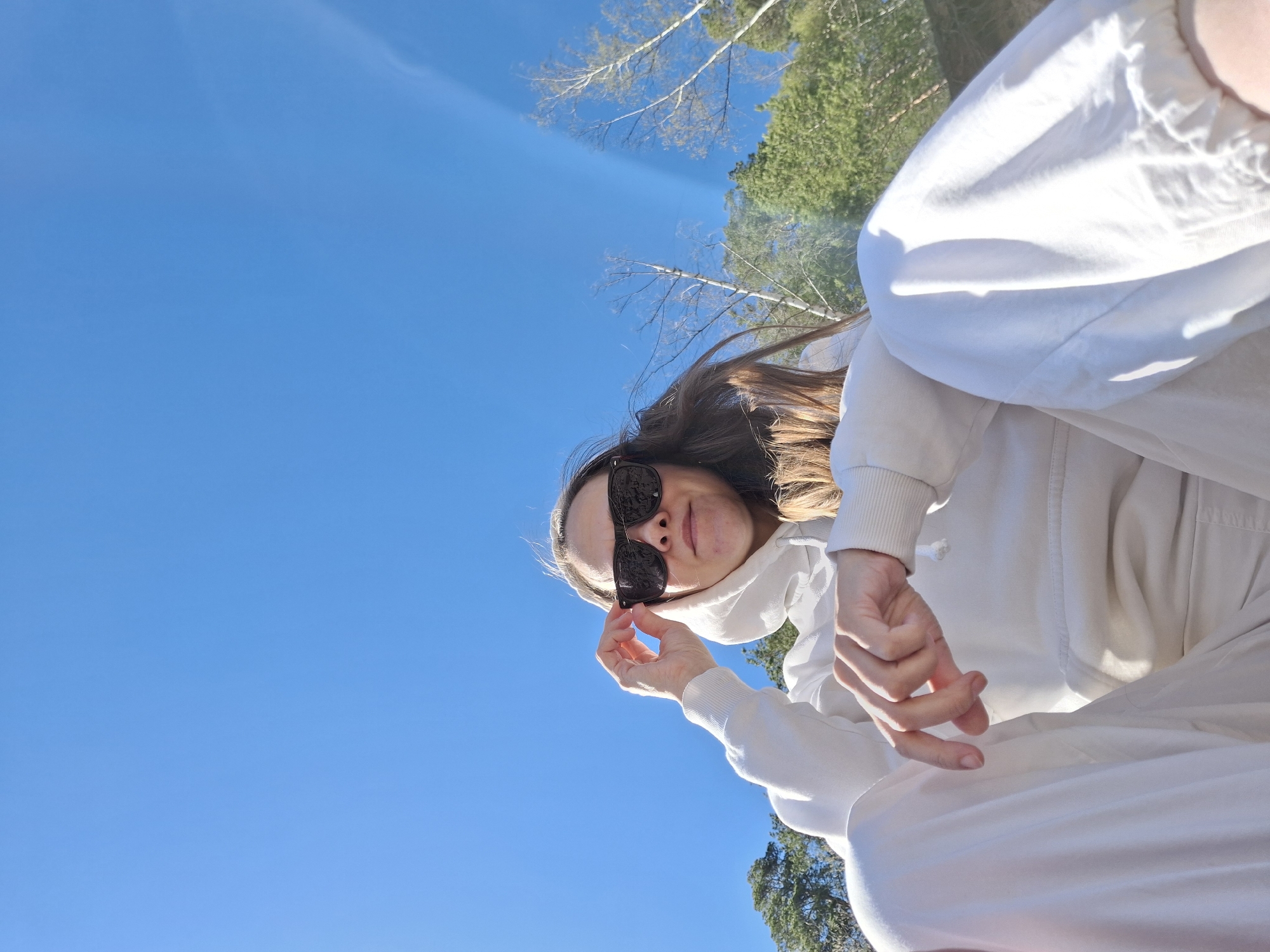 The image is a low-angle selfie taken outdoors in what appears to be a natural setting, possibly near or within the city of Chaikovsky, Russia. The sky is a clear, bright blue. The main subject is a person wearing sunglasses and a white hoodie with matching pants. They are holding the sunglasses, partially obscuring their face. A patch of trees is visible at the top and bottom of the frame, suggesting a park or forest area. The overall impression is a sunny day spent in nature.