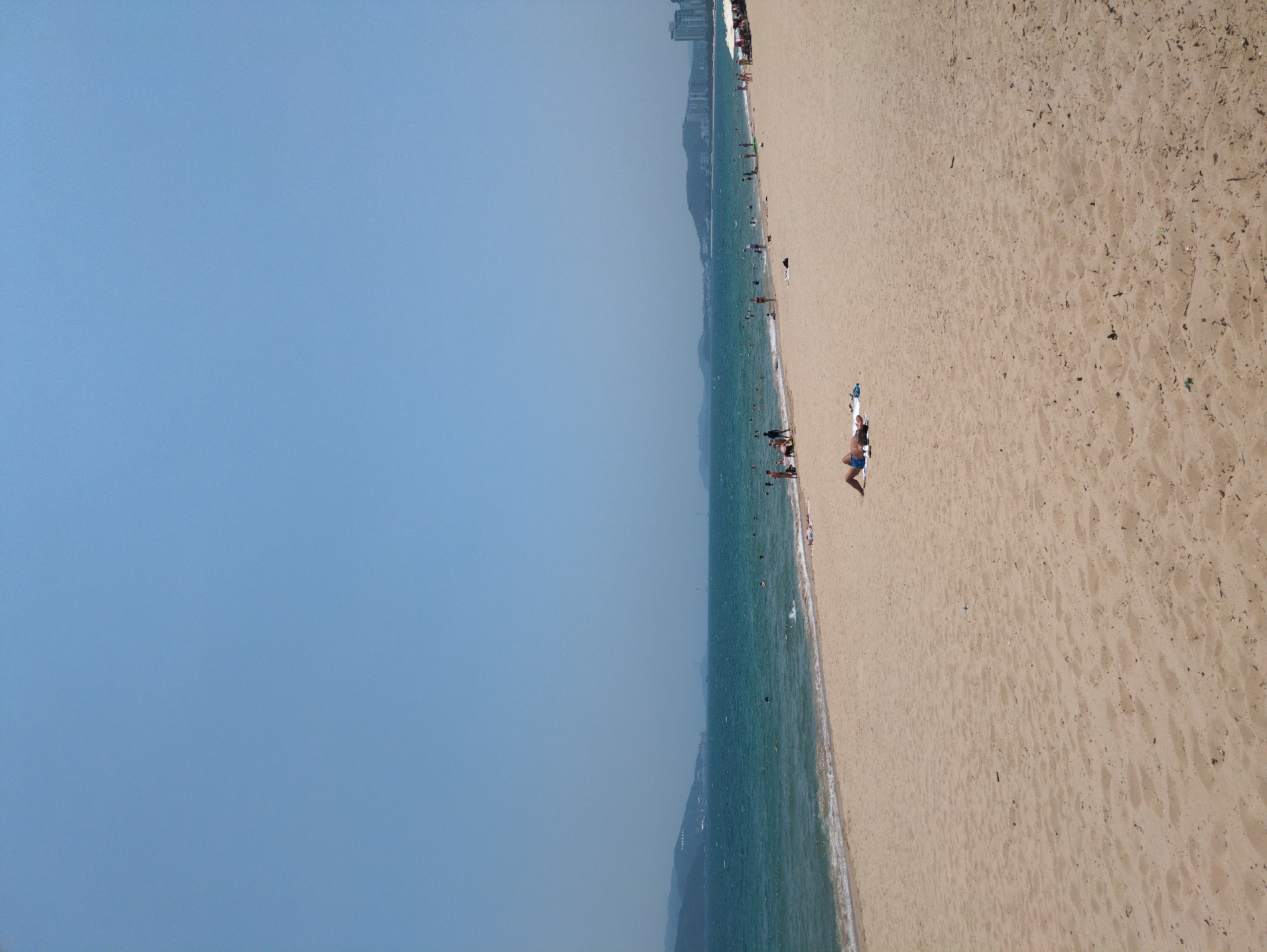 The image shows a beach in Nha Trang, Vietnam. The scene is viewed from an elevated perspective, likely from a high-rise building or elevated viewpoint. The beach is sandy and populated with people engaging in various activities. Some are sunbathing on the sand, while others are swimming in the ocean. A few buildings are visible in the distance along the coastline. The sky is clear, and the sea appears turquoise.