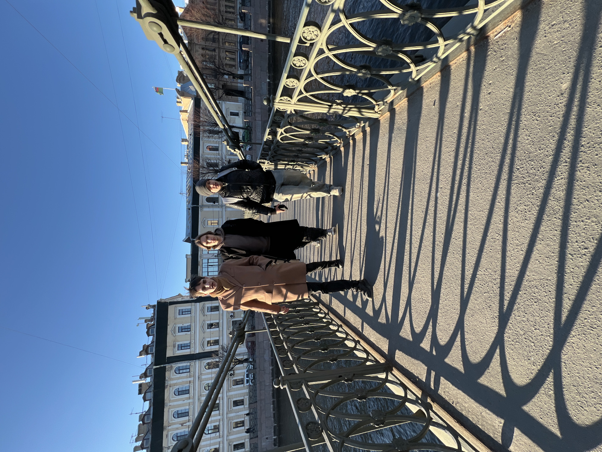 Three individuals stand on a bridge with ornate metal railings casting shadows on the pavement. To the left, a building with multiple windows is visible, along with a glimpse of water. In the background, a flag flies atop a building. The sky is clear and blue. Based on the architectural style and the bridge's design, the location is likely in Saint Petersburg, Russia.
