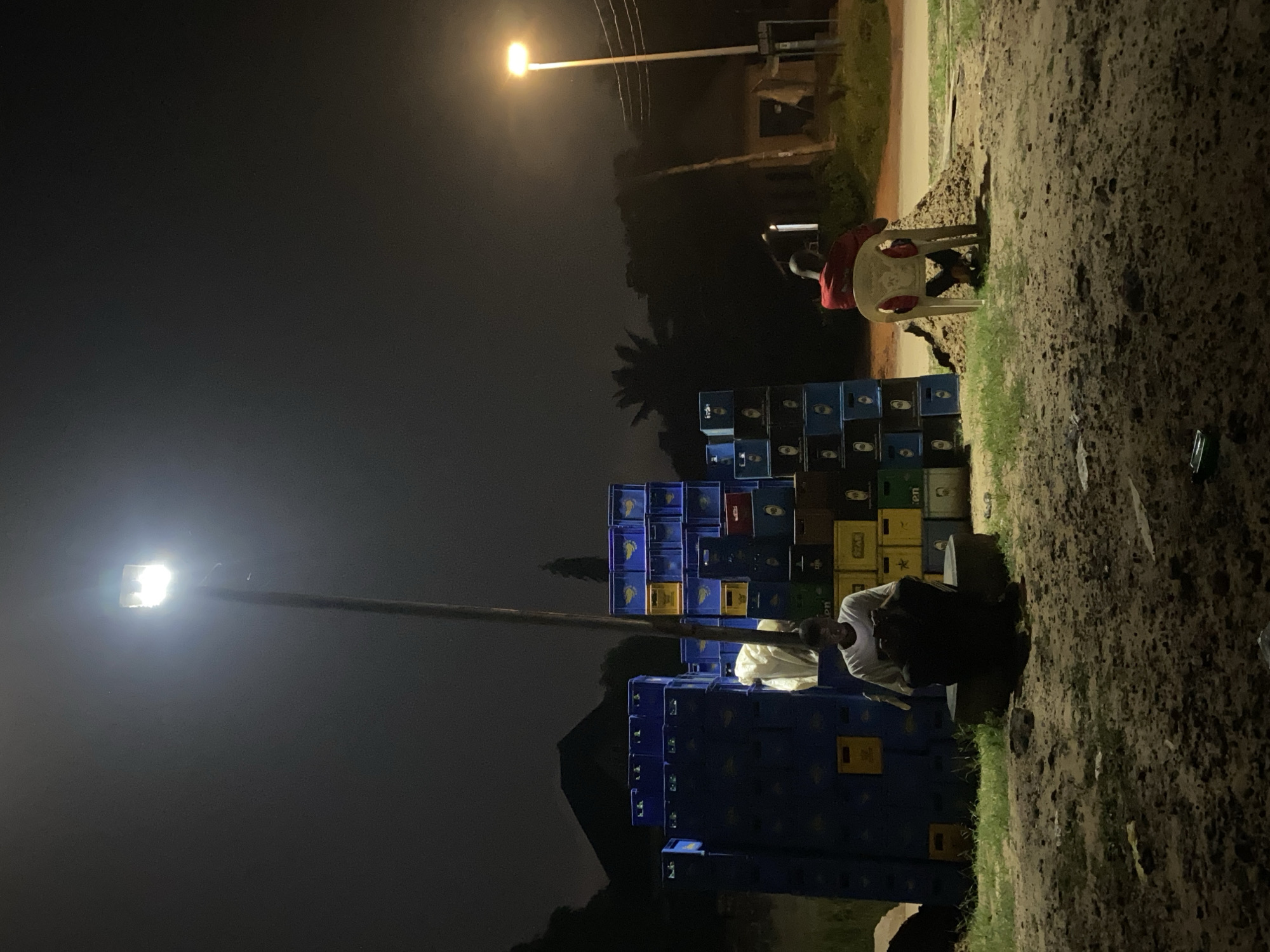 The image depicts a nighttime scene in Nguzu Edda, Nigeria. A large stack of blue, green, yellow, and black beer crates is centrally located. To the right, a person sits on a white plastic chair wearing a red shirt. Another person is squatting near the crates, dressed in a white t-shirt. Two street lights illuminate the area. The ground is dirt with patches of grass.