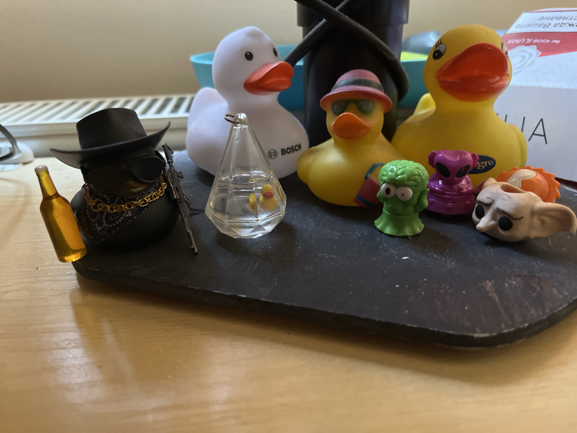 The image shows a collection of novelty rubber ducks and small figurines arranged on a dark rectangular surface. On the left, a black rubber duck wears a cowboy hat, sunglasses, a gold chain, and a bandana, and holds a miniature bottle and rifle. Next to it, a clear glass ornament contains two tiny yellow rubber ducks. A white rubber duck with orange beak and the Bosch logo is to the right of the ornament. A yellow rubber duck wearing a pink hat and sunglasses is positioned in front of the white duck. Further to the right are a green alien figurine, a purple alien figurine, and a light-colored figurine with large eyes. In the background, a red and white box is partially visible. The scene is set on a wooden surface. The image does not contain any information to indicate its location in Sofia, Bulgaria.