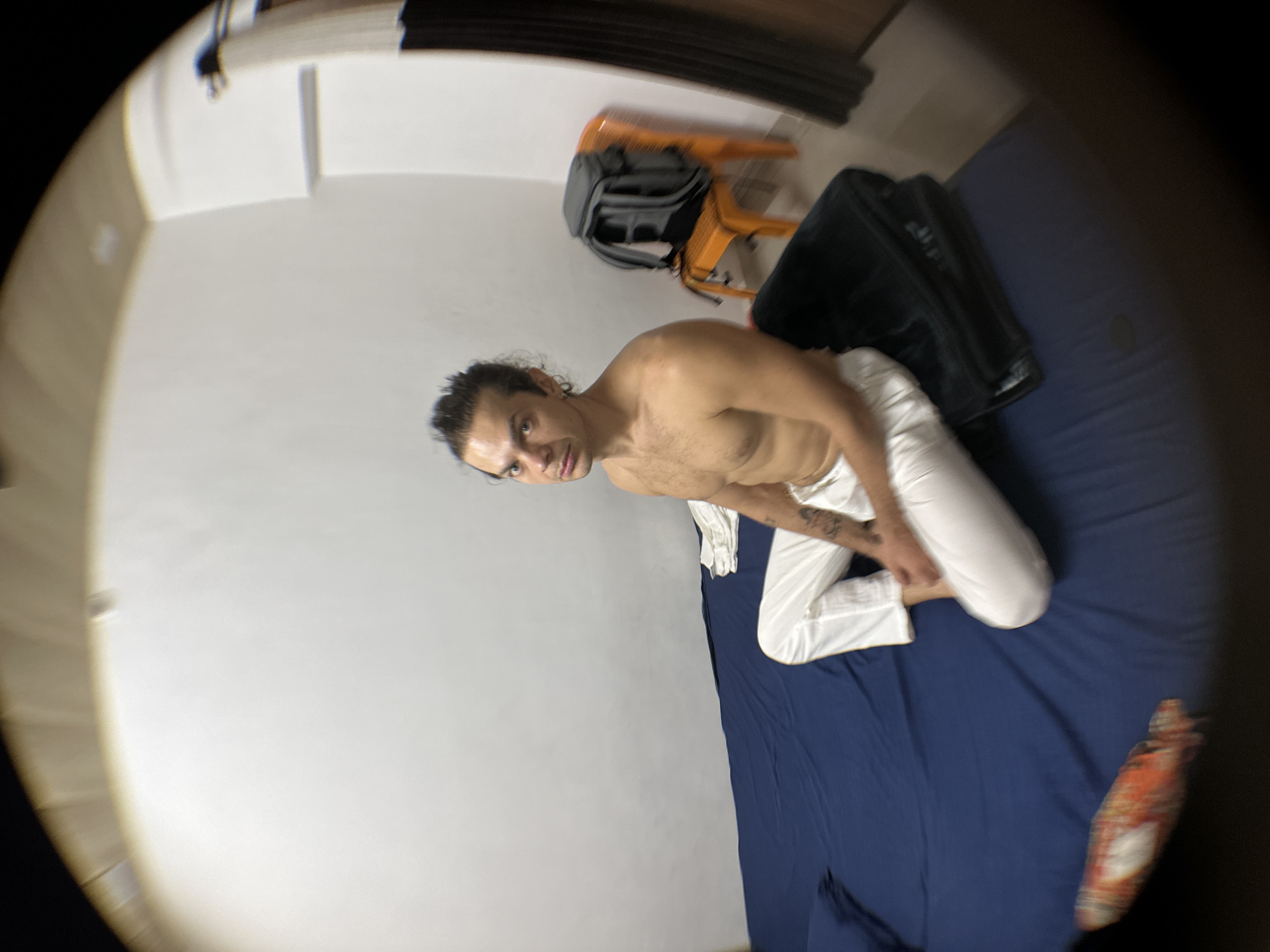 A shirtless man wearing white pants kneels on a blue bedspread in an indoor space. A backpack and black bag are present on a chair in the background. The location is identified as Mathura, India.