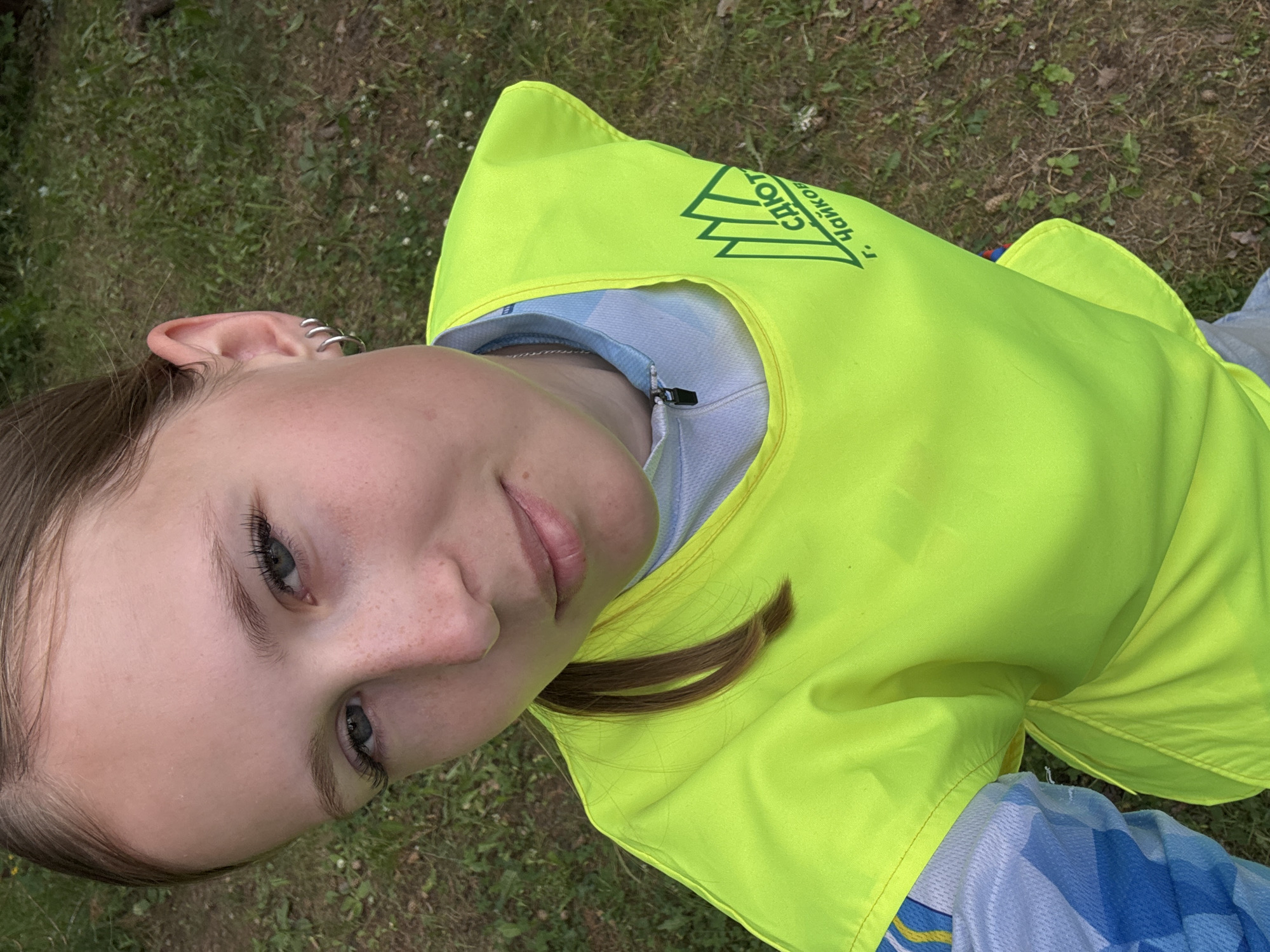 This is a close-up, overhead shot of a young person wearing a bright yellow safety vest over a blue and white athletic shirt. The person has fair skin with freckles and light blue eyes. They have on multiple silver hoop earrings in their left ear. Their hair is brown and is partially visible around their face and shoulder.
The person appears to be lying down or positioned in a way that their face is angled upwards towards the camera. The background consists of green grass with some small white flowers and fallen leaves, suggesting an outdoor setting. The lighting indicates it is daytime, likely sunny, as there are no harsh shadows and the colors are vibrant.
On the yellow vest, there is visible text and a logo. The text appears to be in Russian, and the logo is a green triangle with stylized mountain or abstract shapes inside. The visible text includes "САИОТ" and "г. Чайков".
The overall mood of the image is casual and relaxed. The focus is on the person's face and attire, with a slight tilt to the camera's perspective. The "г. Чайков" likely refers to the city of Chaikovsky in Russia, providing a specific location context.