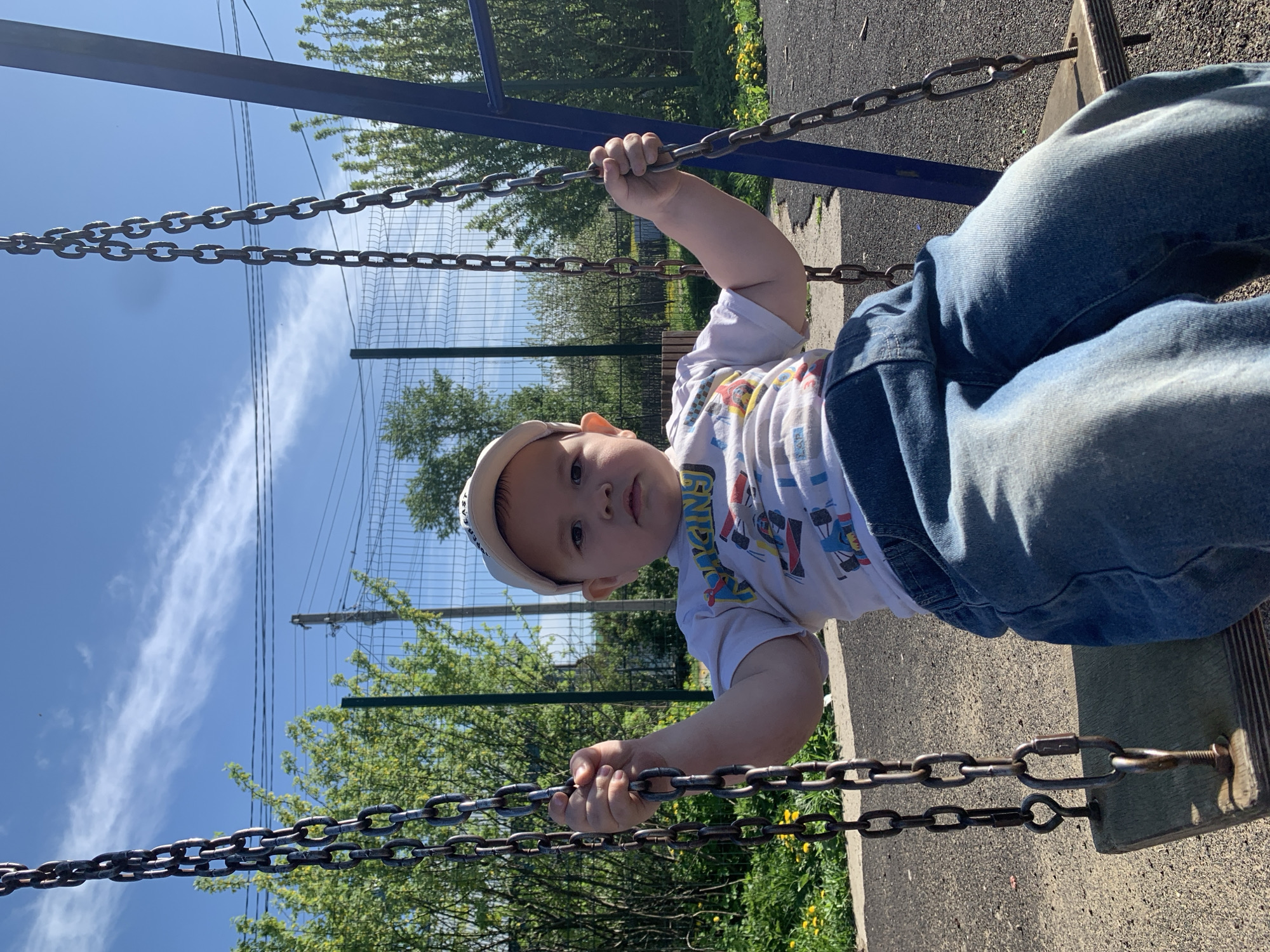 A child is seated on a swing set in a location with trees, a fence, and a blue sky with clouds in the background. The location may be in the city of Фоки, Russia. The child is wearing a white t-shirt with designs, jeans, and a light-colored cap. They are holding onto the swing's chains.