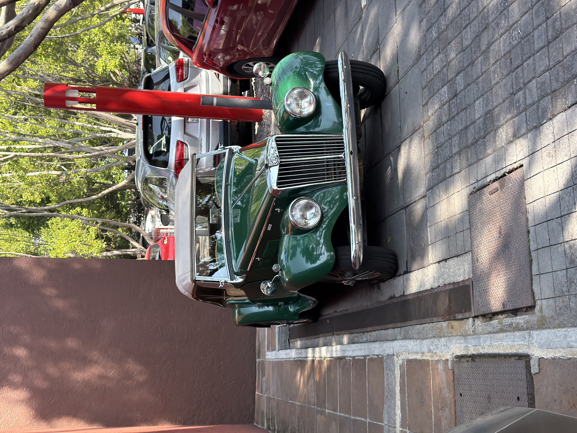 A vibrant green vintage MG car is parked on a gray brick sidewalk next to a brown wall. The car faces the viewer, its chrome grill and headlights prominently displayed. Behind the MG, a red bollard stands near several parked modern cars, including a red SUV and silver vehicles. Trees with green foliage are visible in the background. The scene is captured on a bright, sunny day. A storm drain and a manhole cover are visible near the parked MG car. The location is Puebla, Mexico.