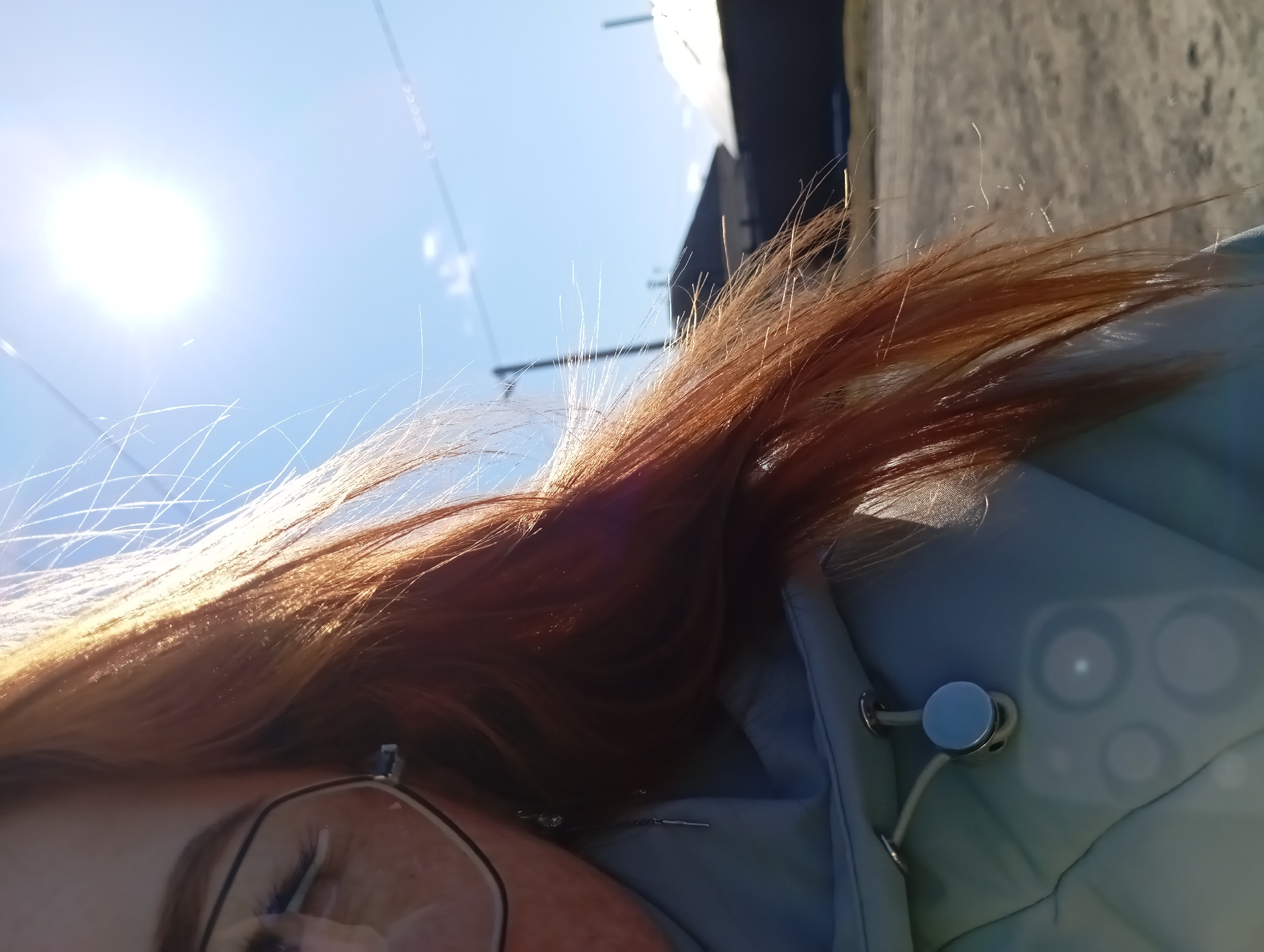 The image shows a close-up, tilted view of a person outdoors in Yurgamysh, Russia. The sun is shining brightly in a blue sky. Part of the person's face, including an eye with glasses, is visible. They have red-brown hair that is partially covering their face. The person is wearing a light blue jacket. In the background, there is a building. Overhead wires are also visible against the sky.