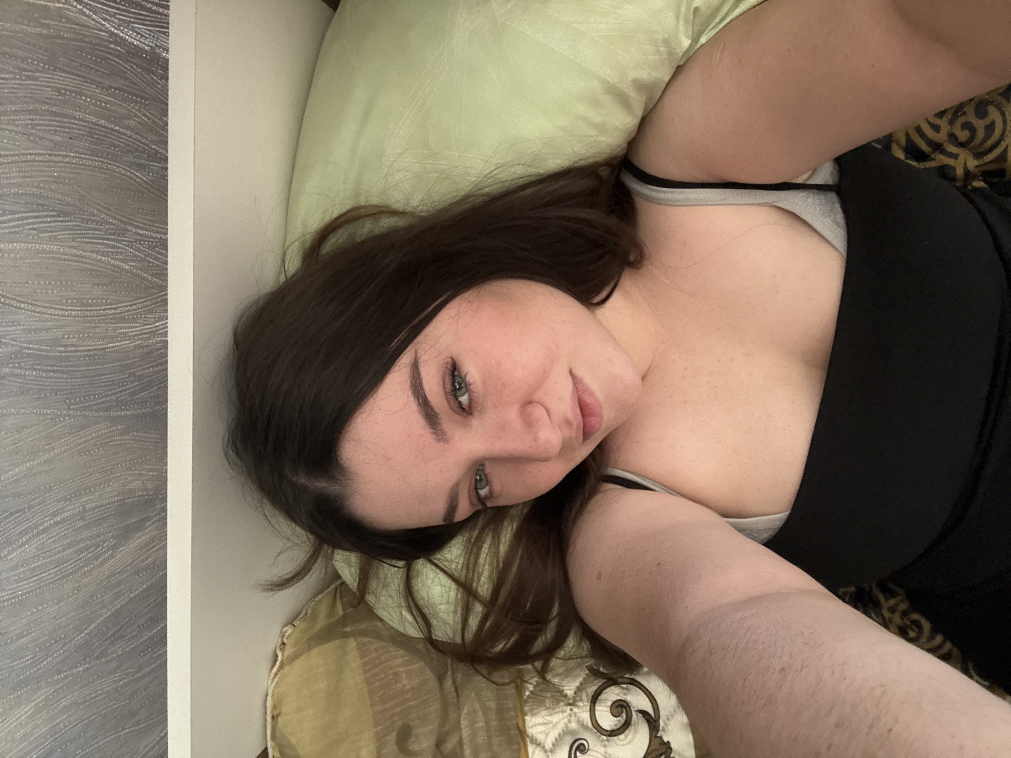 A young woman with long, dark brown hair and light skin is lying on her back in a bed. She is looking up at the camera and smiling slightly. She is wearing a black tank top over a gray bra. Her left arm is raised, and her hand is holding the camera. The background shows a light green pillow and a patterned bedspread. The room appears to be dimly lit, and the overall atmosphere is relaxed and intimate. The image was taken in Borodulino, Russia.