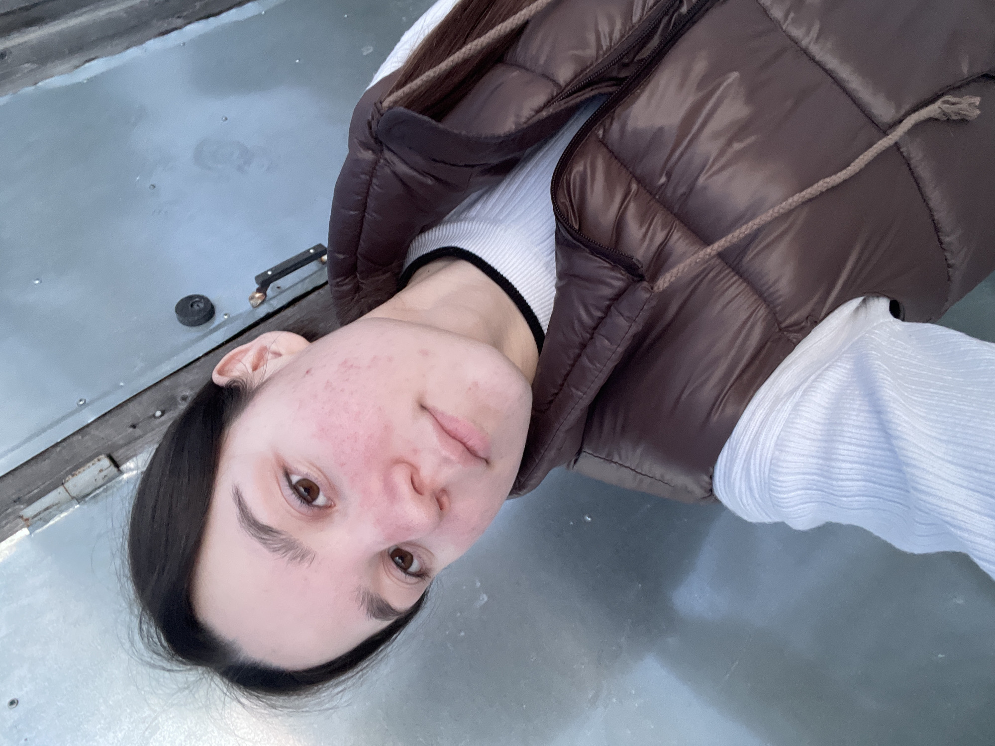 A young woman wearing a white top and a brown padded vest is lying on a gray surface. The surface appears to be outdoors, possibly metallic and reflective. The photo is taken from above, looking down at the woman. The location is Ольховка, Russia.