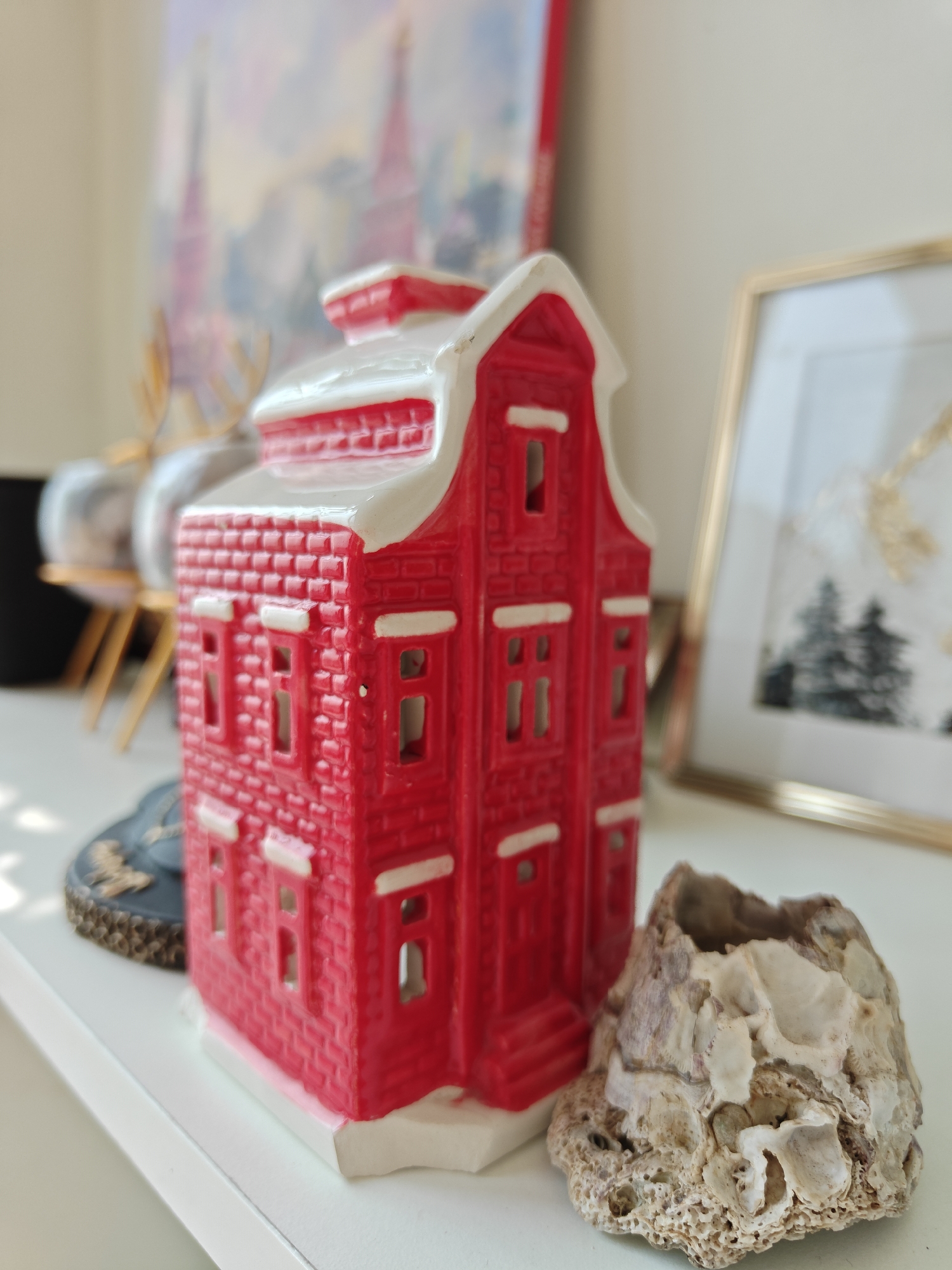 A close-up shot shows a red ceramic house with white trim, likely a decorative object. The house has a brick-like texture, visible windows, and a stylized roof. It sits on a white surface, presumably a shelf. To the right of the house is a natural object appearing to be a weathered rock or a piece of coral. Behind the house, slightly out of focus, are other decorative items: a framed picture with gold trim and an artwork (or a print) with a depiction of winter trees and a painting of what might be the Kremlin in the background. A black object and a clear glass sphere (likely a vase or decorative ball) on a gold stand are seen on the left. There are no people visible. The location cannot be determined from the image, and there is no evidence to suggest the picture was taken in Mumbai, India.