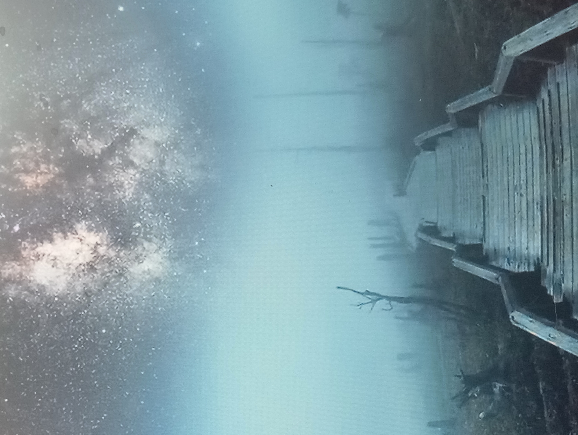 The image shows a wooden boardwalk extending into a body of water, possibly a lake or marsh, with the Milky Way galaxy visible in the sky above. The boardwalk is constructed of weathered planks and has railings on one side. The water is still and reflects the sky. The scene suggests a remote or natural location. There is no information to support any location in Karaganda, Kazakhstan. The image does not contain any identifiable people, specific objects, or actions beyond the static scene. The environment is a combination of water, wooden structure, and celestial display.