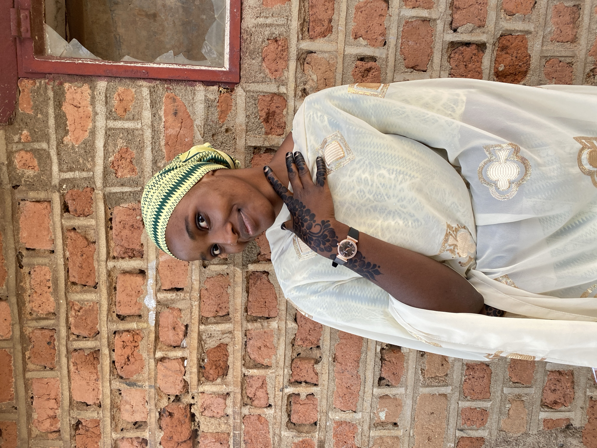 A young woman is seen in front of a brick wall with a red-framed window. The glass pane of the window is broken. She is wearing a green and yellow head covering and a white dress adorned with gold decorations. The woman's right hand is decorated with a dark floral henna design, and she is wearing a watch on her left wrist. Her fingernails are painted black. She is smiling slightly and looking at the camera. The location is Vom, Nigeria.