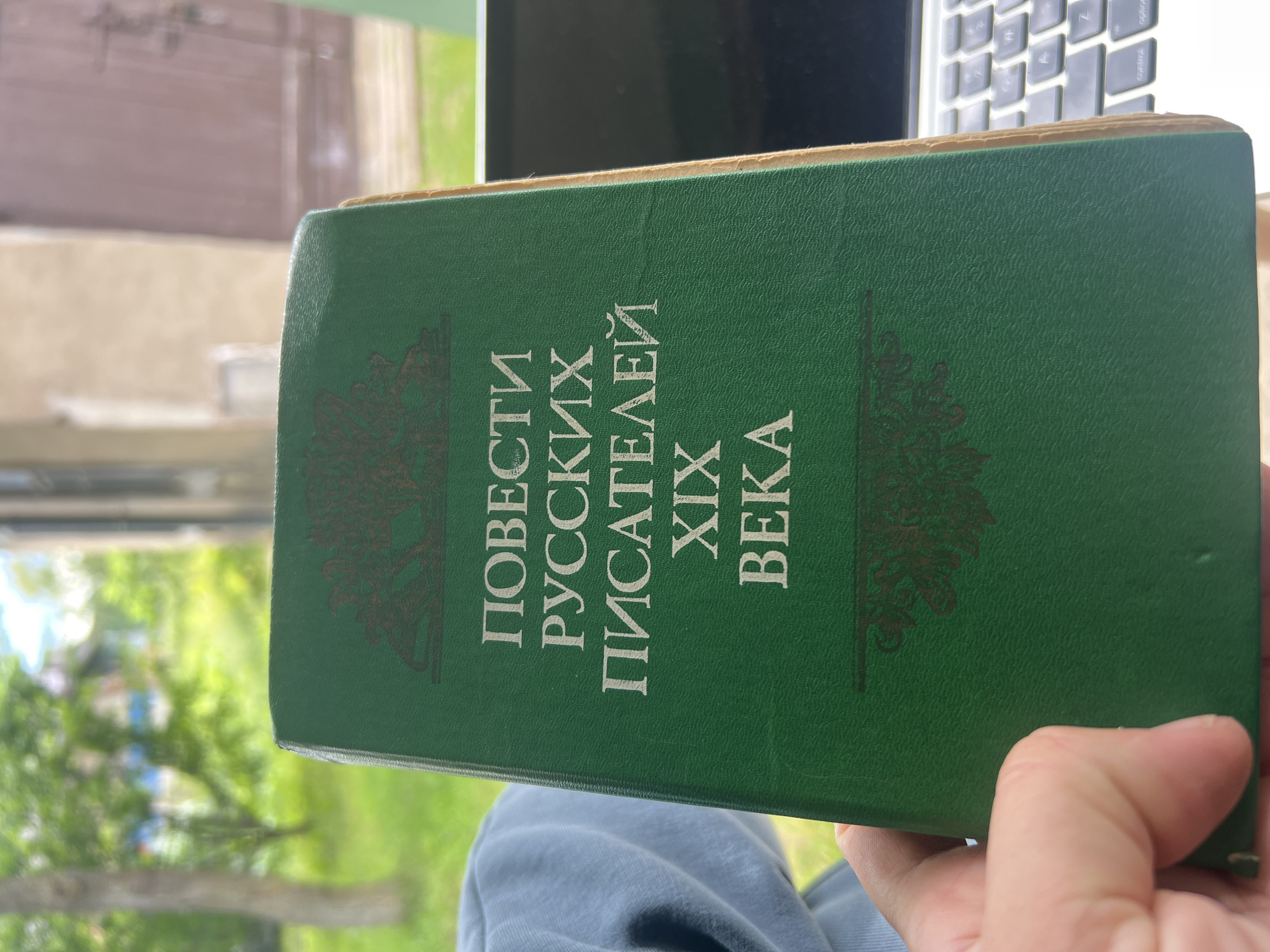 A green book with gold lettering in Russian is held by a person's left hand. The lettering on the cover reads "ПОВЕСТИ РУССКИХ ПИСАТЕЛЕЙ XIX ВЕКА," which translates to "Stories of Russian Writers of the 19th Century." The book is positioned in the foreground, obscuring the lower portion of a laptop keyboard. The background is blurred, showing a bright, grassy outdoor environment with trees, suggesting a sunny day. The image appears to be taken from a slightly elevated angle, looking down at the book. The location of Бучацінскі сельскі Савет, Belarus is not discernible from the visual information provided in the image.