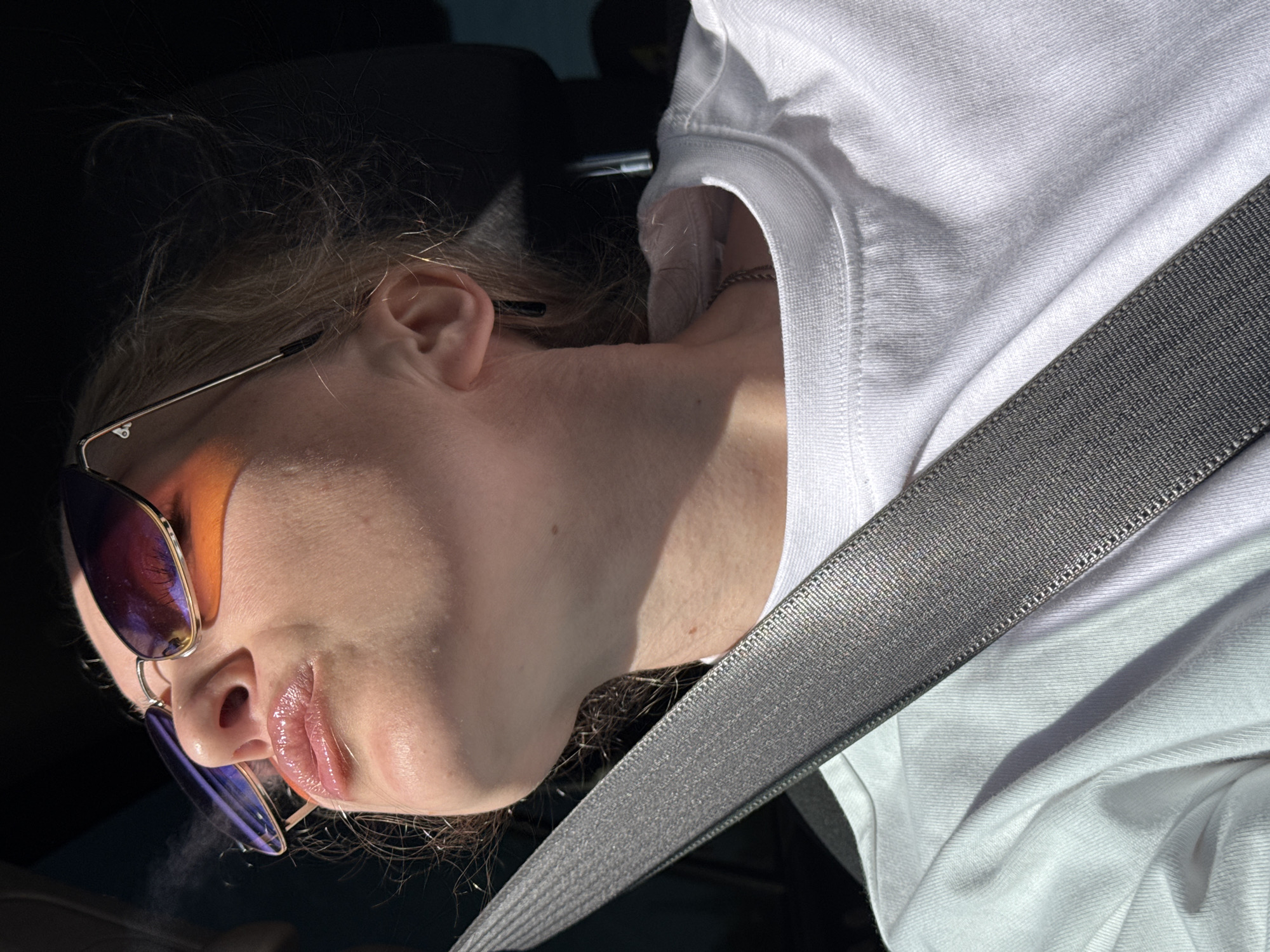 A close-up, angled shot depicts a person with light skin wearing sunglasses, a white T-shirt, and a seatbelt, suggesting they are in a vehicle. Wisps of what appears to be vapor or smoke emanate from near the lips. The lighting is uneven, with strong shadows and bright sunlight illuminating parts of the face and clothing. The exact location within the city of Chaikovsky, Russia, cannot be determined from the image alone. There is no further information available.