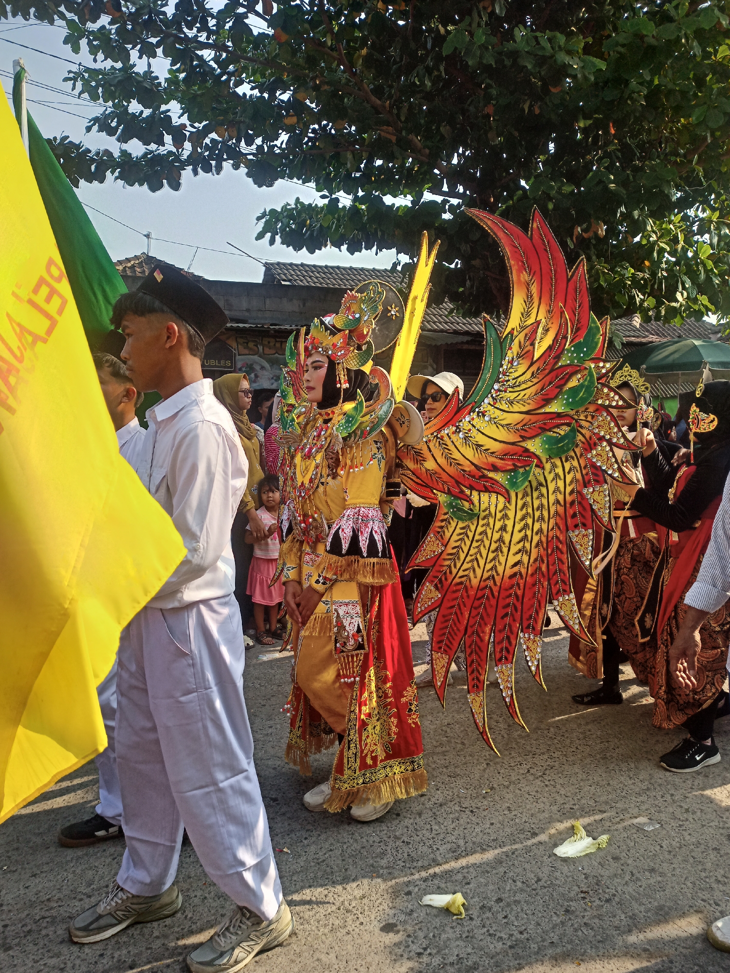 In Saren, Indonesia, a procession is underway on a paved road, with people participating and observing. The central figure is adorned in a vibrant, yellow and red costume featuring elaborate wings resembling those of a mythical bird. The costume is richly decorated with gold trim and beaded embellishments. To the left, a young person wearing a black cap and white long-sleeved shirt holds a large yellow flag. Behind them, other individuals are visible, some wearing traditional Indonesian attire such as batik sarongs. A young child in a pink dress stands among the onlookers. Trees with green foliage line the background, and buildings with tiled roofs are visible. The scene is lit by natural daylight, casting shadows on the ground.