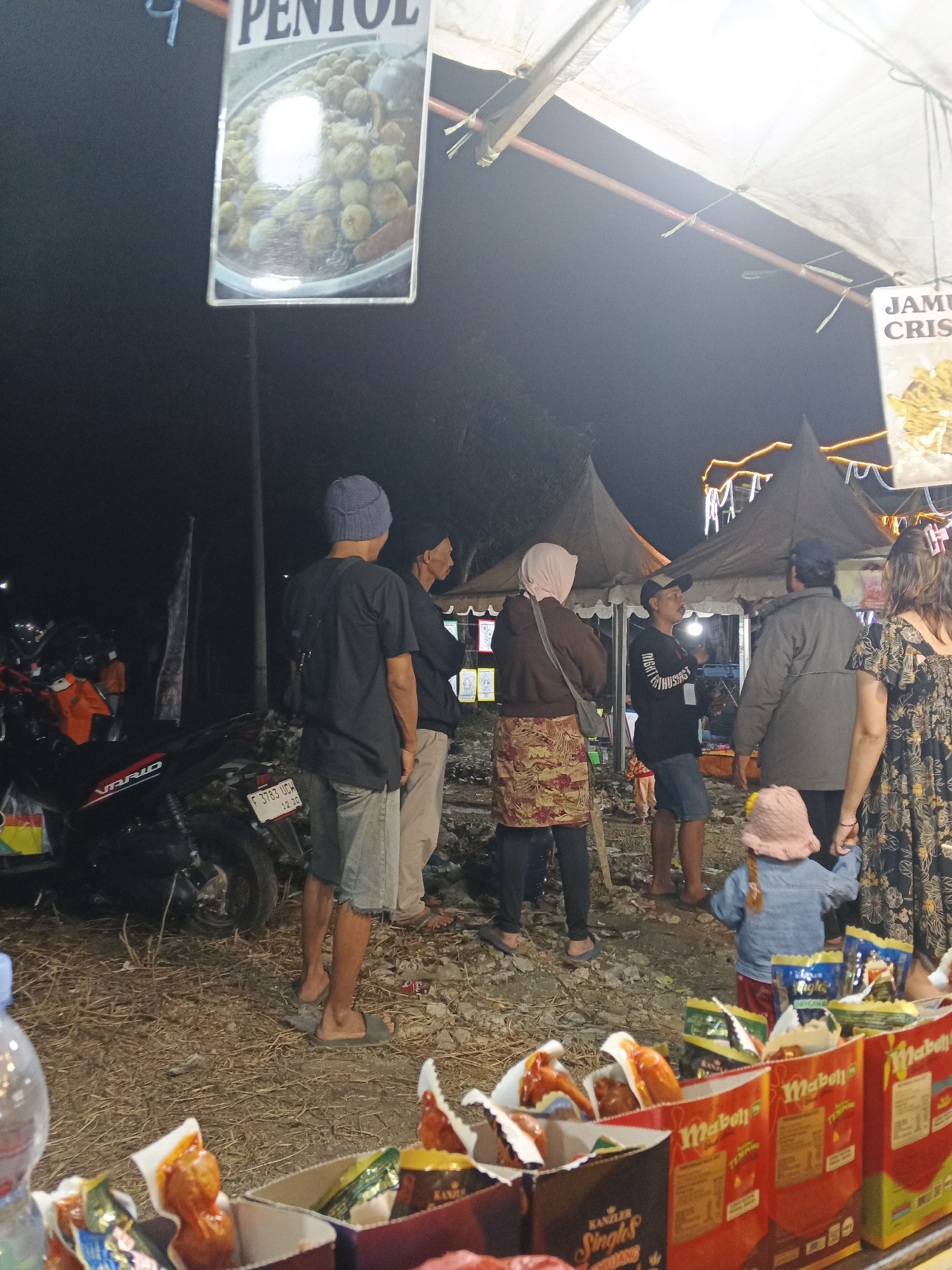 The image depicts a nighttime scene at what appears to be a street food market in Sukabumi, Indonesia. In the foreground, several boxes of packaged food products are displayed. Behind these boxes, a group of people are gathered. A man wearing a blue beanie and a dark t-shirt stands with his back to the camera, facing another man in a dark jacket. To their left, a woman in a brown hoodie and a patterned skirt is visible, and a young child wearing a pink hat and a denim jacket is being held by a woman in a floral dress. Several tents are set up in the background, illuminated by string lights. To the far left, a motorcycle is parked. The ground is covered with a mixture of dry straw and debris. Two signs are visible; one at the top left has the word "PENTOL" and an image of small, round food items, while another sign on the right has the text "JAMU CR..." with an image of crunchy snacks. The overall atmosphere suggests a local gathering or festival.