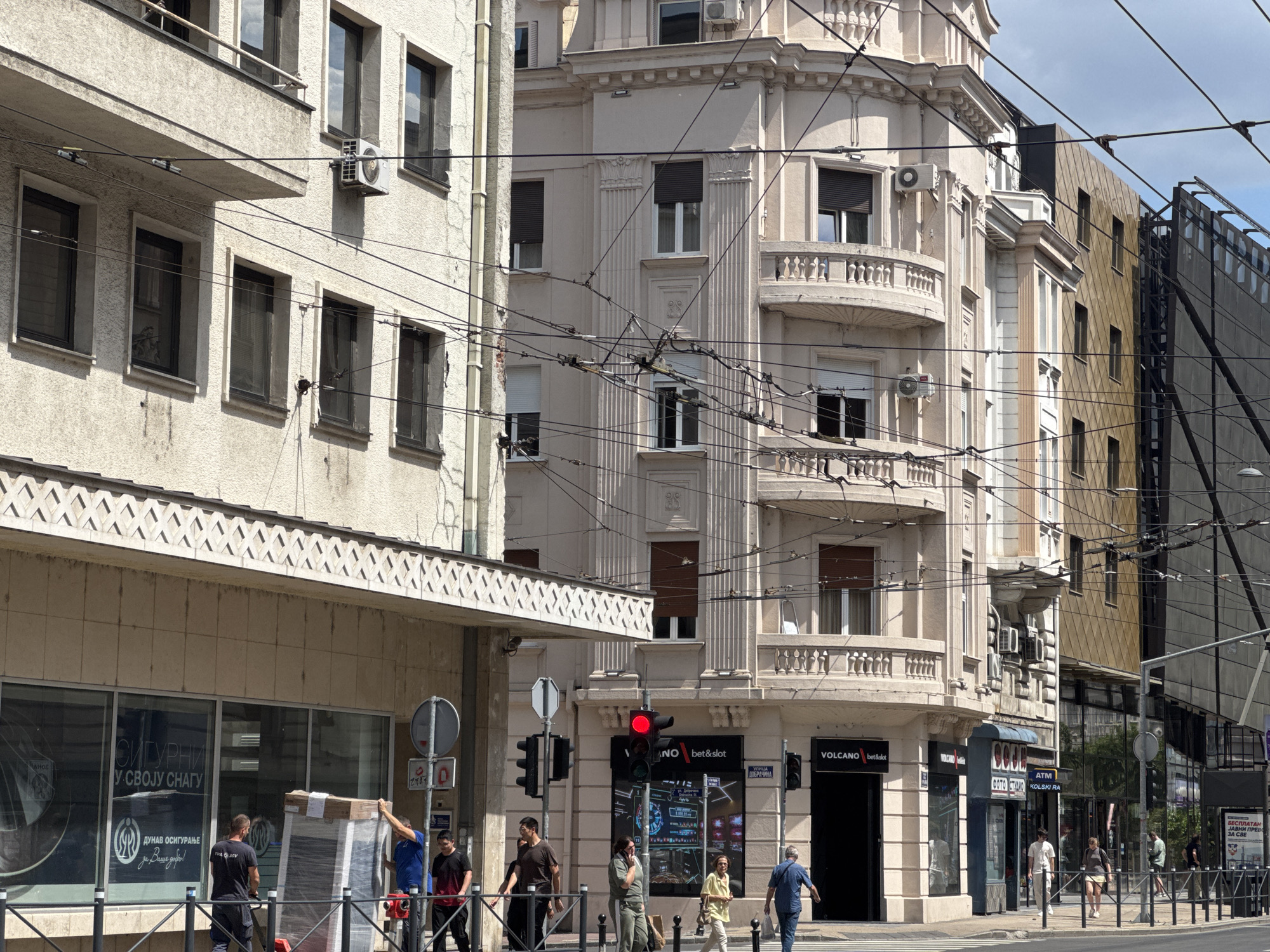 The image depicts a street scene in Belgrade, Serbia, likely in the city center due to the presence of multiple buildings with commercial establishments and a busy pedestrian area. The architecture suggests a mix of older, ornate European style buildings and more modern ones. The street is lined with power lines and trolleybus wires, indicating public transportation is active in the area.
In the foreground, several people are visible. On the left, three men are engaged in moving a large, wrapped object, possibly a piece of furniture or appliance, which is secured with plastic wrap. One man wears a dark t-shirt with "VOLCANO" printed on the back, suggesting a connection to a nearby establishment. To the right of the moving men, pedestrians are walking along the sidewalk. Further to the right, a traffic light shows red, and a sign for "VOLCANO bet&slot" is prominently displayed on a building.
The buildings in the background are multi-story structures. One building, painted a light pink, features decorative elements, balconies, and numerous windows. Another building to its right has a facade with a geometric, diamond-patterned texture and reflective windows. The presence of outdoor air conditioning units on some buildings is noticeable. The street appears to be paved, and there are metal railings along the sidewalk. The sky is partly cloudy.