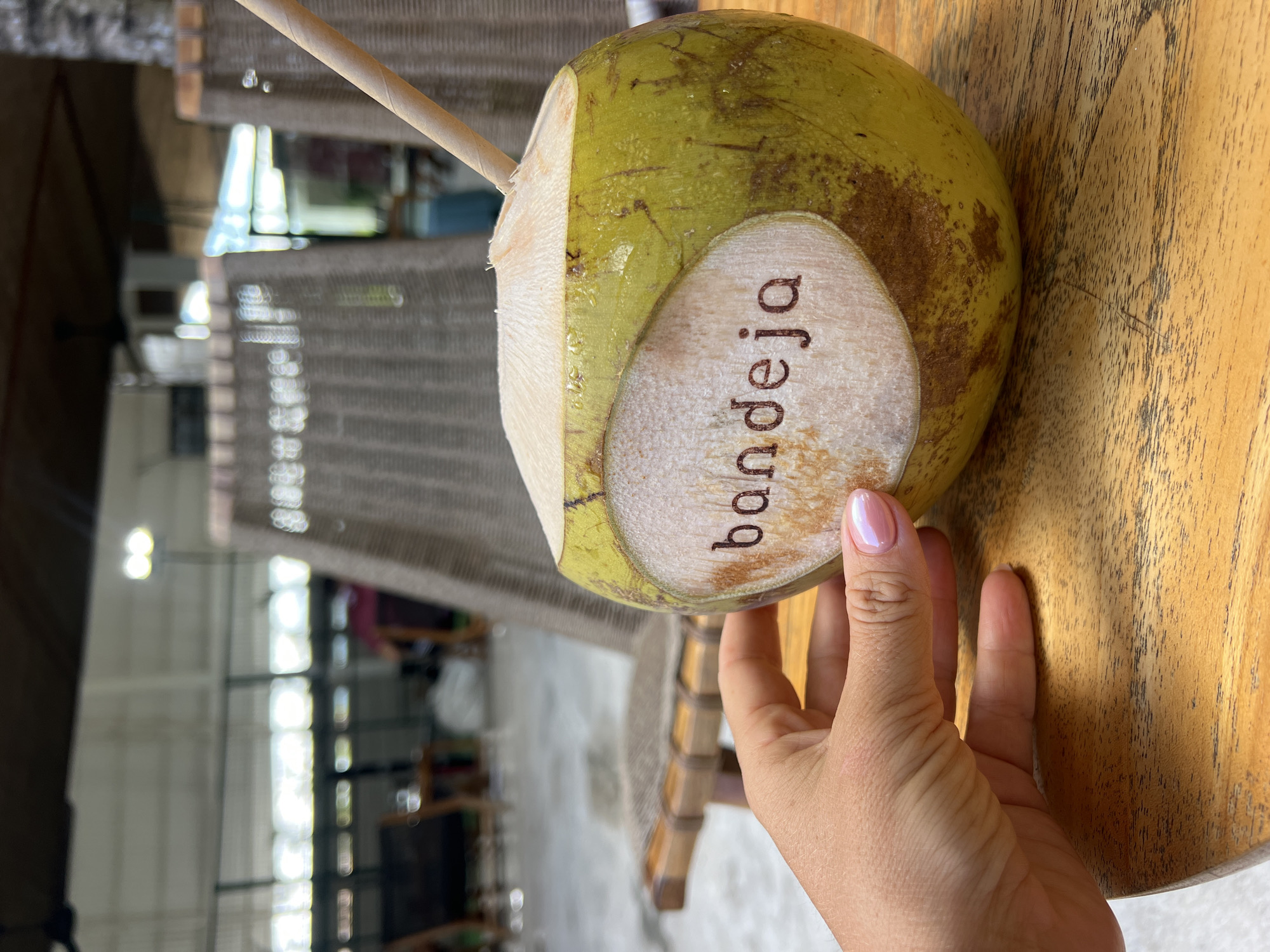 A close-up, eye-level shot shows a person's left hand gently holding a young, green coconut with a paper straw inserted into the top. The coconut has a light brown oval label with the word "bandeja" engraved on it. The coconut rests on a wooden table. In the background, wicker chairs and a blurred outdoor seating area are visible, suggesting a casual outdoor establishment. The scene is lit by natural daylight, contributing to a relaxed atmosphere. The location is identified as North Kuta, Indonesia.