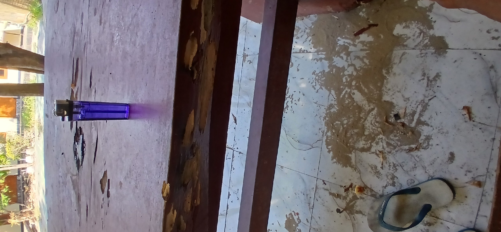 The image depicts a purple lighter standing upright on a weathered wooden table. The table's surface shows signs of damage and wear. Below the table, the tiled floor is scattered with debris, leaves, and what appears to be a wet substance. A light-colored flip-flop with a dark strap rests on the floor. In the background, there are glimpses of vegetation, trees, and buildings, suggesting an outdoor or semi-outdoor setting. The scene appears to be from a residential area in Pejaten, Indonesia, possibly a veranda or patio, given the presence of the table, floor, and open-air elements. There are no people visible in the image.