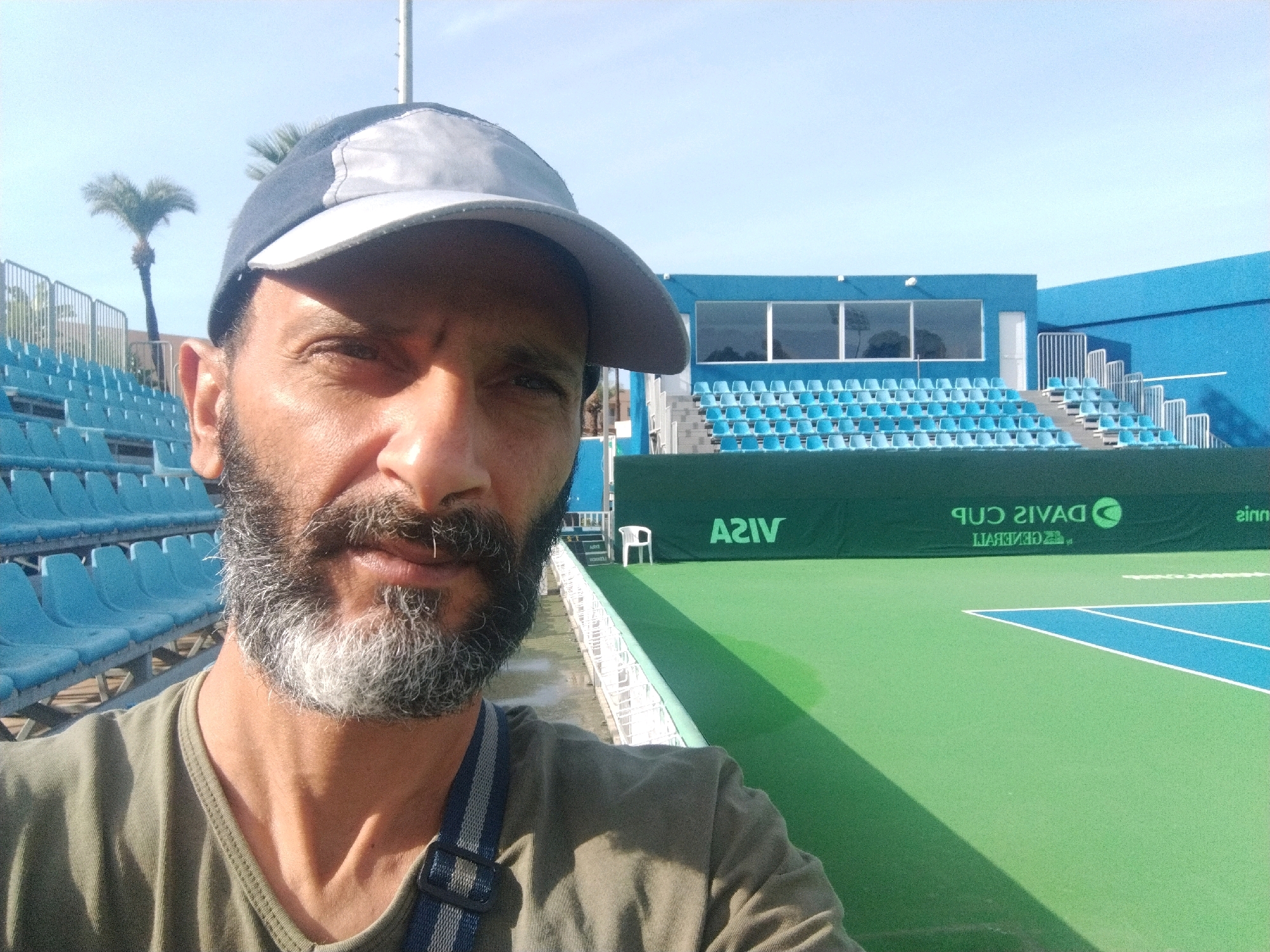 A medium shot photograph shows a man with a beard and a baseball cap taking a selfie outdoors at a tennis court. The man is wearing a green t-shirt and a striped strap across his chest. Behind him, the tennis court is visible with green turf and blue lines. The stands, filled with empty light blue seats, are situated to the left of the court, and a blue building with windows is in the background. A palm tree is visible to the far left, and a green banner with logos is displayed behind the court. The sky is clear and blue. The scene appears to be at the tennis courts in Monastir, Tunisia, possibly during a Davis Cup event, as indicated by the banner which shows the "Davis Cup" logo.