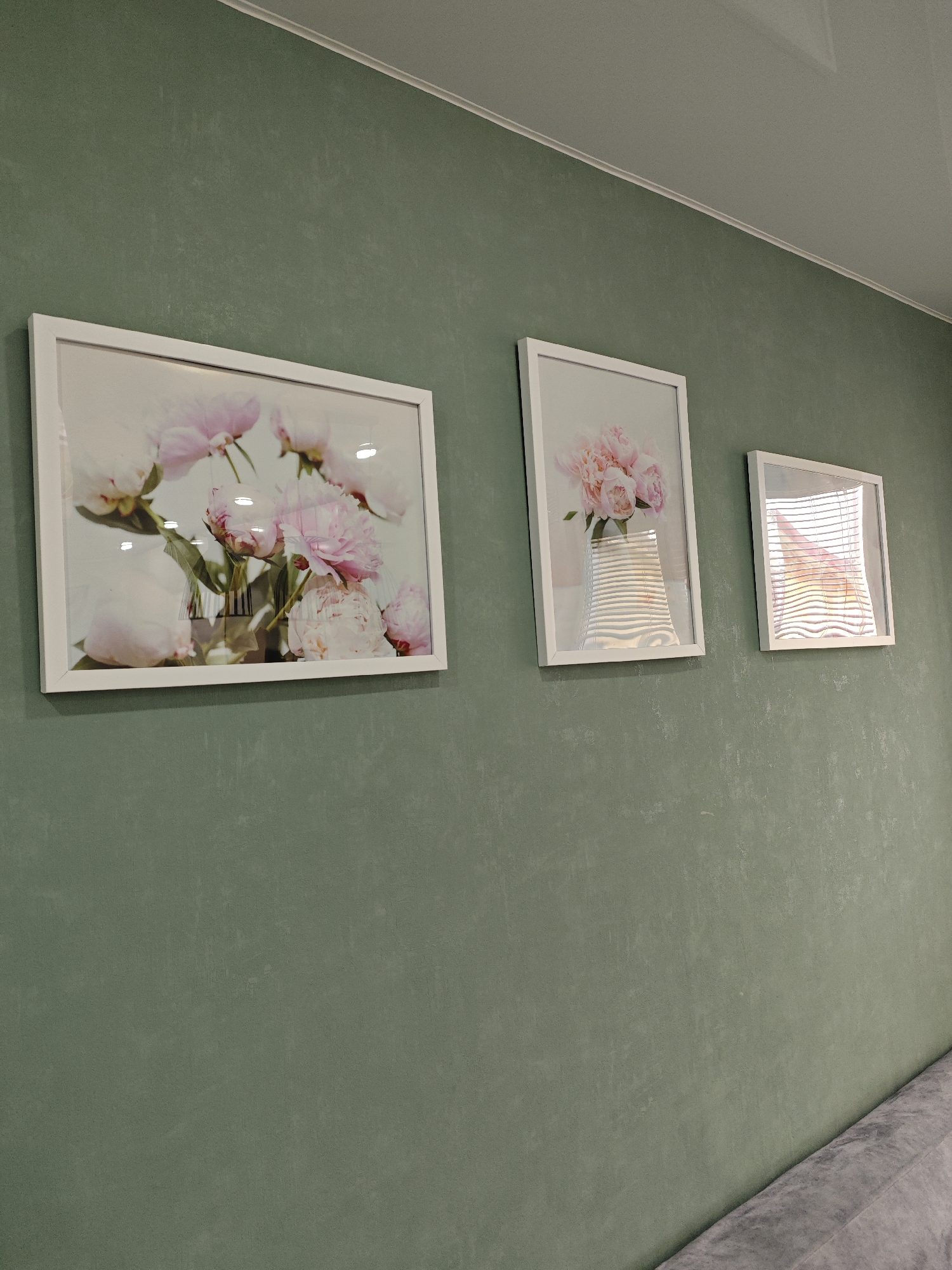 The media file captures an indoor scene, likely within a home or commercial establishment in Фоки, Russia. The dominant feature is a muted green wall, possibly sage green, which exhibits a subtle textured or mottled finish. Along the top, a white crown molding separates the wall from a light-colored ceiling.
Three framed pictures with simple white frames are mounted horizontally on the green wall. They are arranged in descending size from left to right.
The largest picture on the left displays a close-up image of light pink peony flowers in a clear vase, with several blossoms and buds visible. The glass covering the picture shows reflections of overhead light sources.
The middle picture, smaller than the first, also features pink peony flowers in a vase, depicted from a slightly different perspective. This picture's glass also has noticeable reflections, including distinct horizontal lines that suggest venetian blinds or a similar patterned light source.
The smallest picture on the right shows an image of horizontal blinds with an orange-yellow glow emanating from behind them, possibly depicting a sunset or a warm artificial light source through a window.
A hint of a grey, textured furniture piece, possibly a sofa armrest or backrest, is visible in the bottom right corner of the frame. There is no discernible activity taking place, and no text is visible in the image. The lighting suggests daytime, given the reflections on the picture frames.