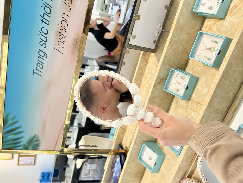A hand holds a seashell-decorated oval mirror, reflecting two kissing people. The mirror's handle is embellished with white seashells. A woman sits in the background. Jewelry displays with light blue boxes line a wall. A sign with Vietnamese text and the words "Fashion Jewelry" is visible to the left.