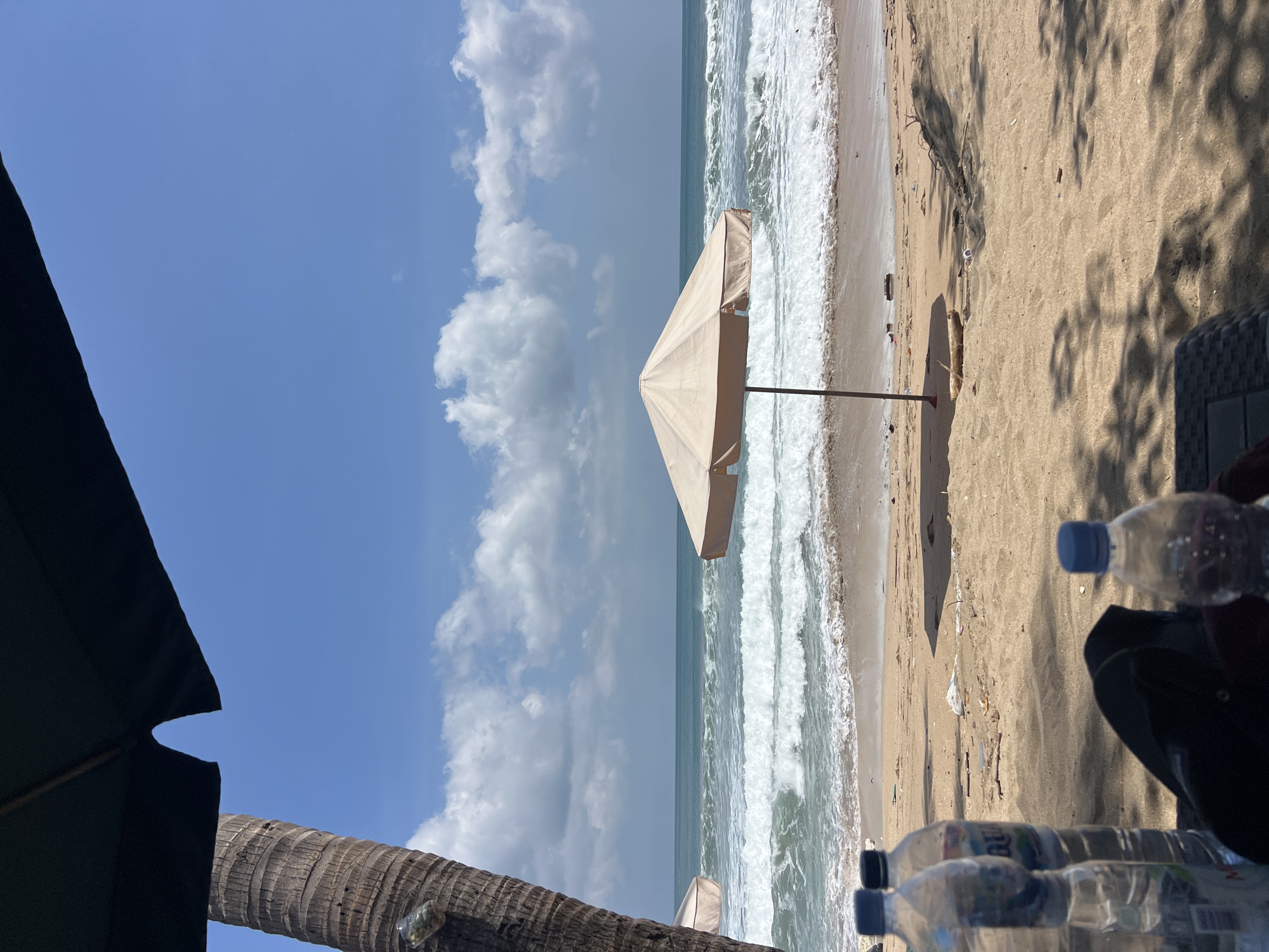 The image depicts a serene beach scene in Kuta, Indonesia, under a bright blue sky with scattered white clouds. In the foreground, several plastic water bottles are visible on the sand, along with a dark object that appears to be a bag or a piece of clothing. A portion of a large palm tree trunk stands to the left, partially obscured by a dark umbrella.
Further back, a light-colored beach umbrella provides shade on the sandy shore. The ocean is calm with gentle waves rolling onto the beach, and the water is a clear turquoise color. The beach itself appears to have some scattered debris, suggesting it may be a public beach. The overall impression is one of a relaxed day at the beach, with elements of nature and human presence coexisting.