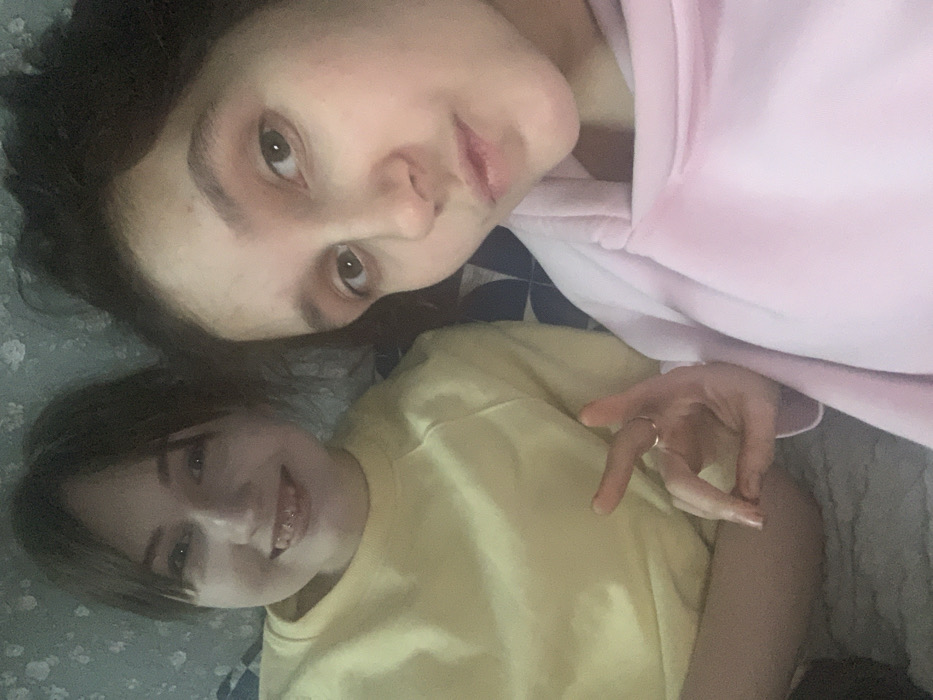 The image depicts two young females in a casual indoor setting within Yekaterinburg, Russia. The precise location within the city is not discernible. The scene appears to be a posed selfie.
The female on the left has light brown hair with bangs and is wearing a light yellow long-sleeved garment. She is smiling, revealing upper teeth fitted with dental braces.
The female on the right, positioned closer to the camera and appearing to be taking the photograph, has darker brown hair and is wearing a light pink hooded sweatshirt. She is looking directly at the camera with a slight pout. Her right hand is visible in the lower center, forming a gesture where the thumb and index finger are connected, and the other three fingers are extended upwards. A thin ring is present on her right ring finger.
The background consists of patterned surfaces: a light-colored wall with a subtle floral or textured pattern behind the female on the left, and a section with blue and white geometric patterns to her right, possibly part of bedding or upholstery. The lighting is consistent with an indoor environment.