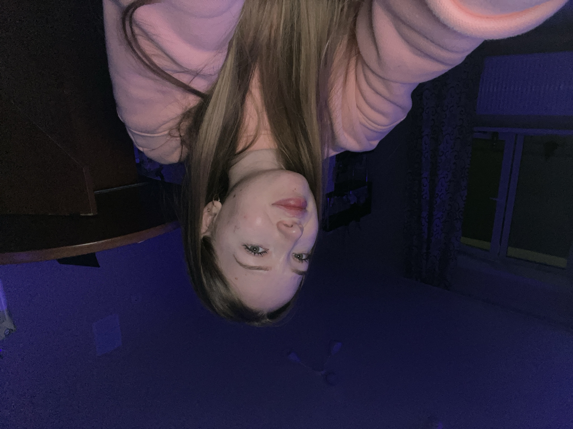 A photograph shows a woman, angled upside down, seemingly taking a selfie indoors. She has long, light brown hair and is wearing a pink hooded sweatshirt. The background includes parts of furniture: a dark wooden table and a window with a patterned curtain. The floor is a dark color, possibly carpeted. The lighting appears to be dim with a bluish cast, giving the scene an overall dark mood. The location is unknown.