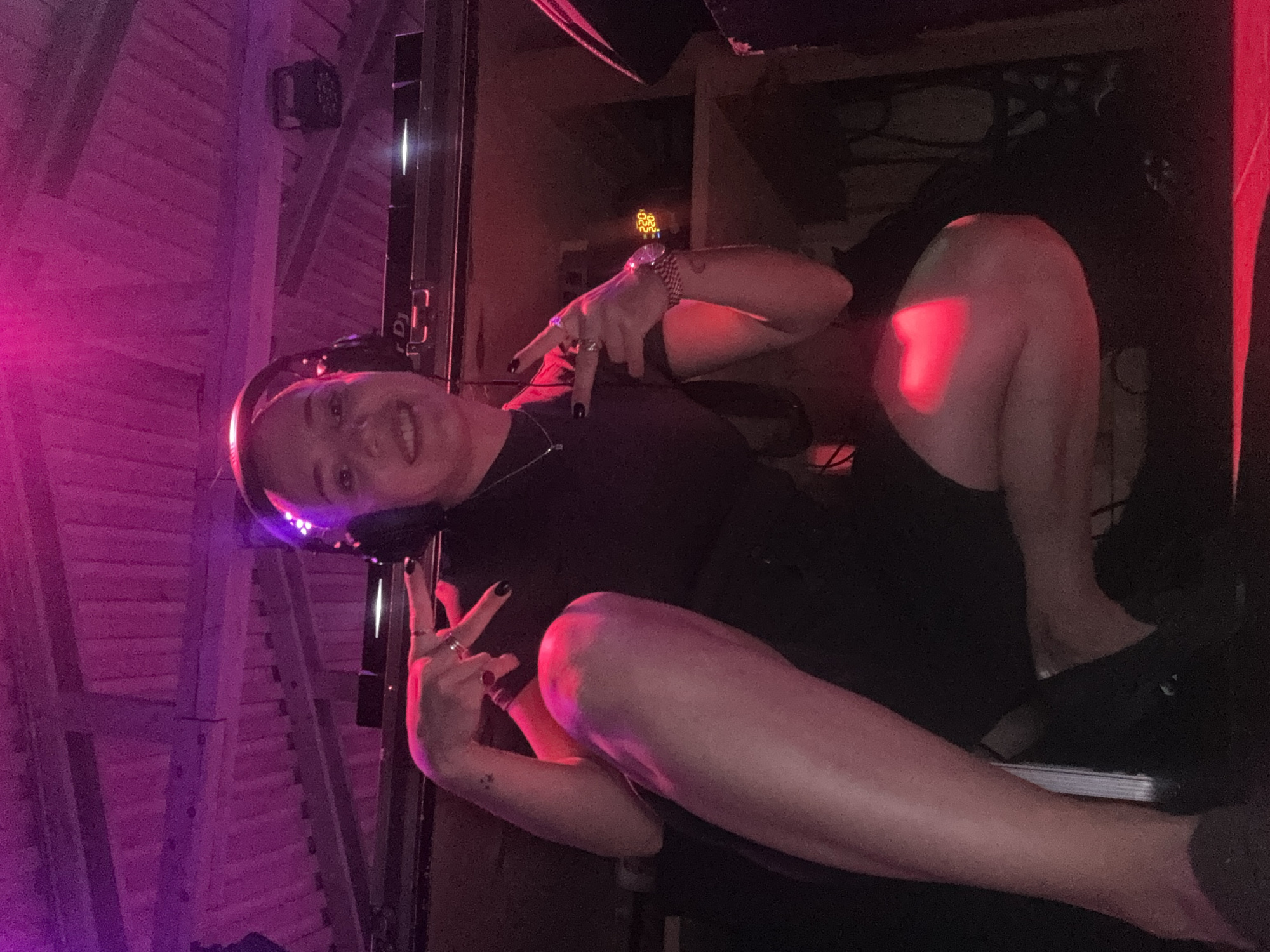 A person with light skin, wearing headphones and black clothing, is seated in a dimly lit room. The person is making peace signs with both hands. The lighting is primarily red and purple, with elements suggesting a music or entertainment venue. The location, Moscow, Russia, cannot be determined from the image.