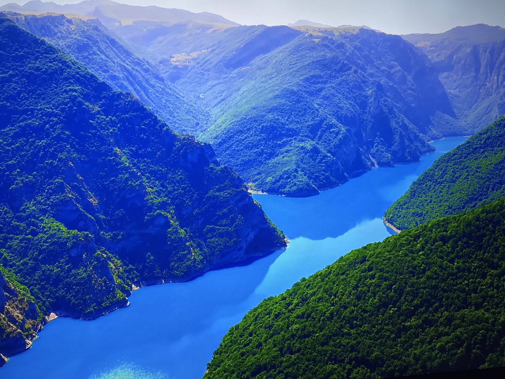This is a high-angle, wide shot of a stunning natural landscape. The dominant feature is a large, winding river or lake with exceptionally vibrant, deep blue water. The water snakes through a canyon-like valley, flanked by steep, verdant mountains covered in lush green trees. The mountains are textured with visible rock formations and slopes. In the distance, the mountains recede into layers of blue and hazy silhouettes under a pale, bright sky. The overall impression is one of vastness and natural beauty. The image does not depict any urban elements or people. The location is not in Burgas, Bulgaria; this is a natural landscape.