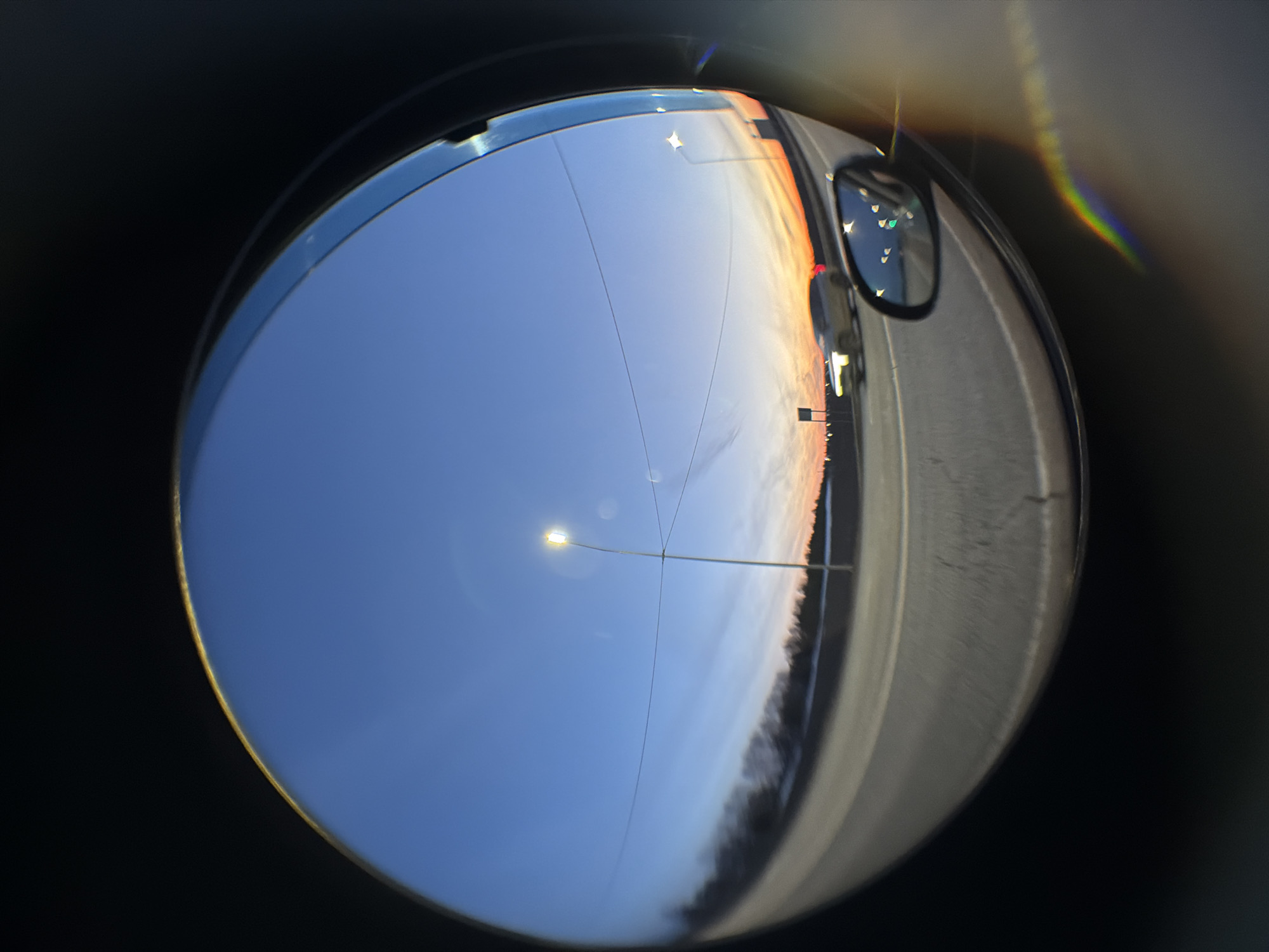 The image depicts a fisheye lens view of an evening scene. A blue sky with a street light dominates the left side of the frame, while a road with a car's side mirror is visible on the right. The horizon shows hues of orange and white, suggesting a sunset or sunrise. A dark line of trees or foliage runs along the bottom of the road. The location in Kuregovo, Russia, cannot be confirmed from the image.