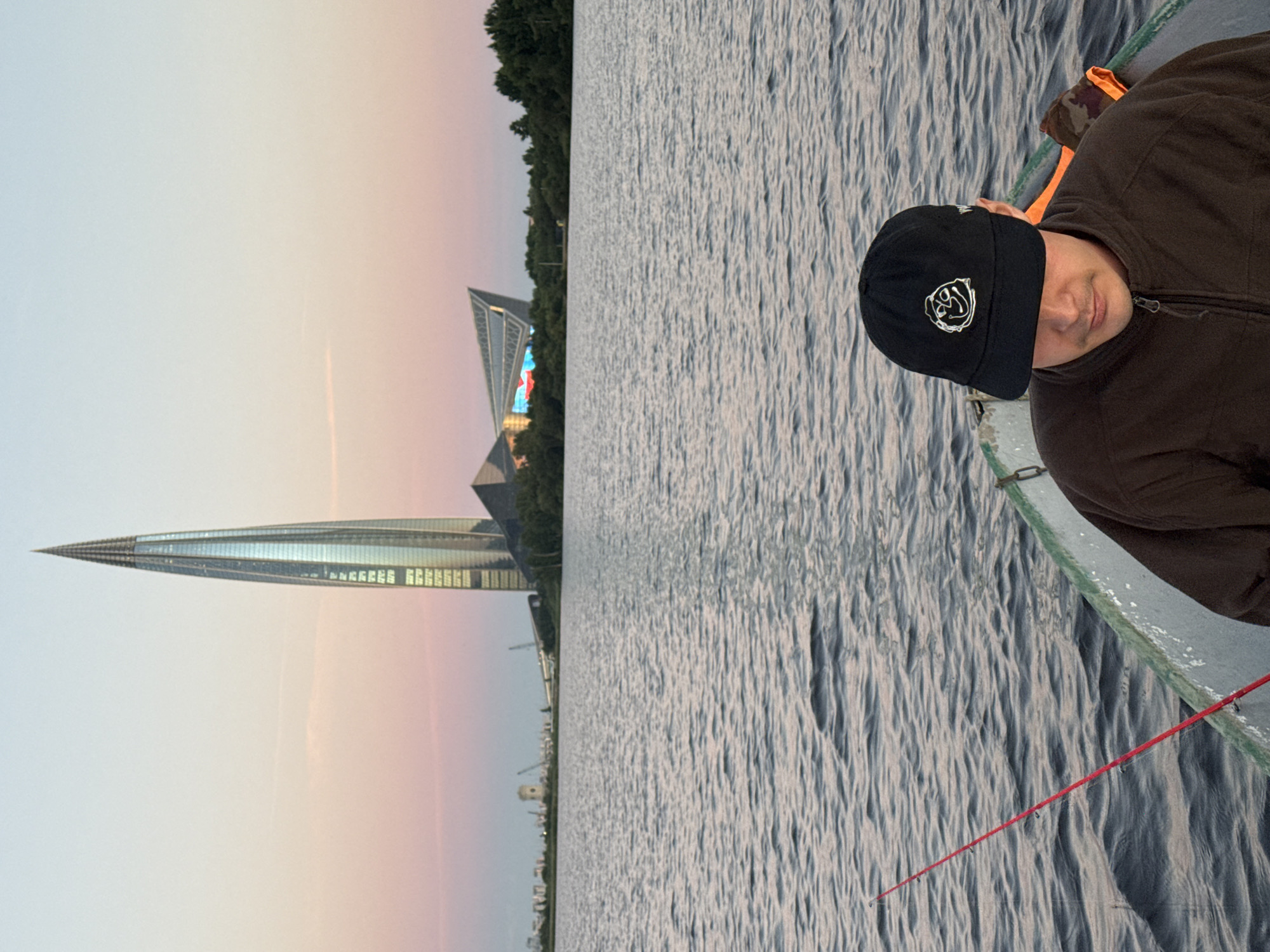 A person wearing a black baseball cap with a cartoon face graphic and a brown zip-up jacket is in a boat on a body of water. A red fishing rod extends from the boat to the left. In the background, across the water, is the cityscape of Saint Petersburg, Russia. Dominating the skyline is the Lakhta Center, a modern skyscraper with a distinctive twisted shape. To its right is a large, angular building that appears to be a stadium or arena, illuminated with colorful lights. The sky above is a gradient of pale blue and pink, suggesting either dawn or dusk. Trees line the shore between the water and the buildings. The water is rippled, indicating a slight breeze. The overall impression is a serene scene of leisure activity with a prominent modern architectural backdrop.