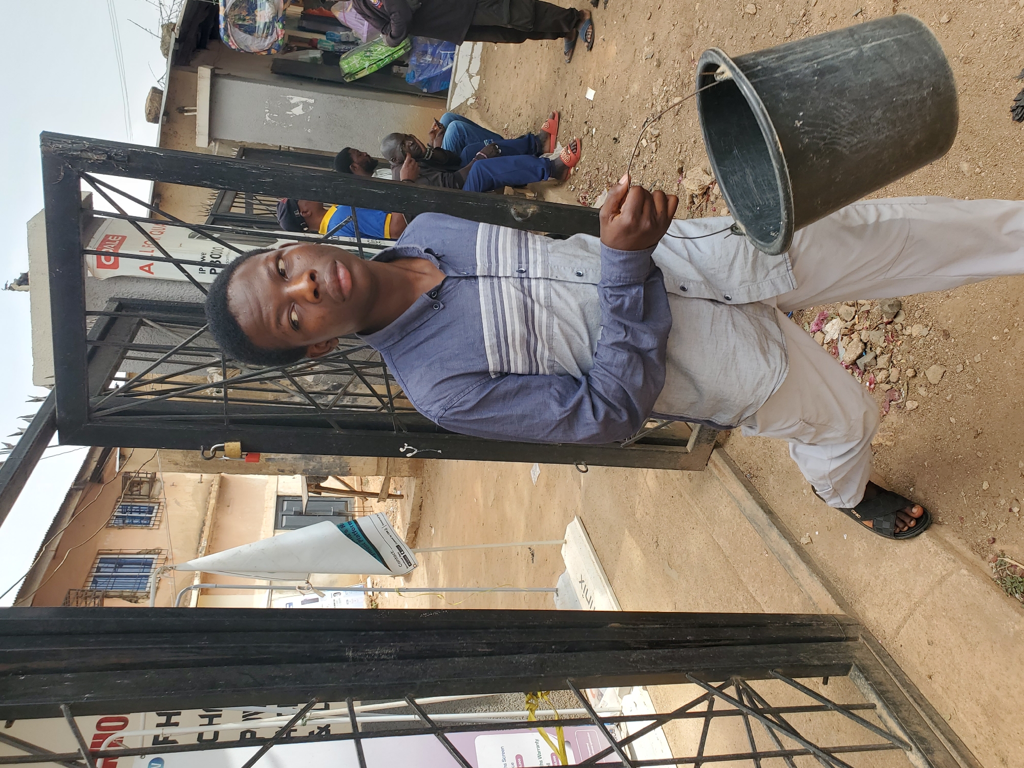 The image shows a man in Jos, Nigeria, holding a black bucket. He is standing in front of a black metal gate or fence. The man is wearing a button-up shirt with gray and blue stripes, light-colored pants, and black sandals. Other people are visible in the background, some sitting and others standing. A building is visible behind the gate, and various signs are present, some with legible text such as "FH", "CH", "inix", "CALE", "IP we", and "P30000". The ground is dirt.