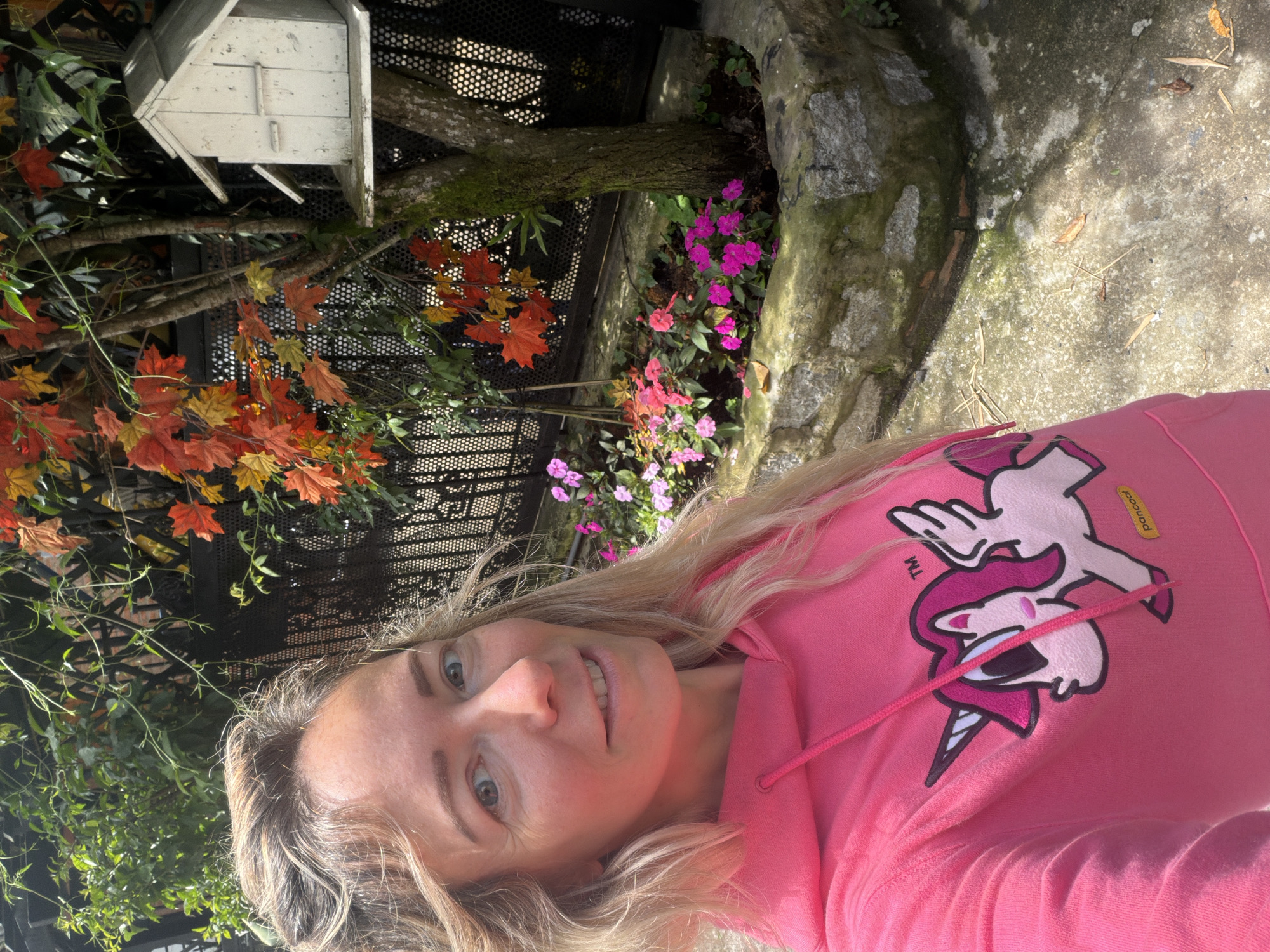 A blonde woman with light blue eyes and fair skin, wearing a bright pink hoodie with a cartoon unicorn graphic, poses for a selfie outdoors. The background features a garden area with a small, weathered white birdhouse mounted on a tree. Red and orange autumn leaves are visible on the tree, contrasting with lush green foliage and vibrant pink and fuchsia flowers planted in a stone-lined bed. The ground appears to be a concrete pathway with scattered dry leaves and twigs. The scene suggests a pleasant day in Da Lat, Vietnam, a city known for its cooler climate and French colonial architecture, often adorned with flowers and trees.