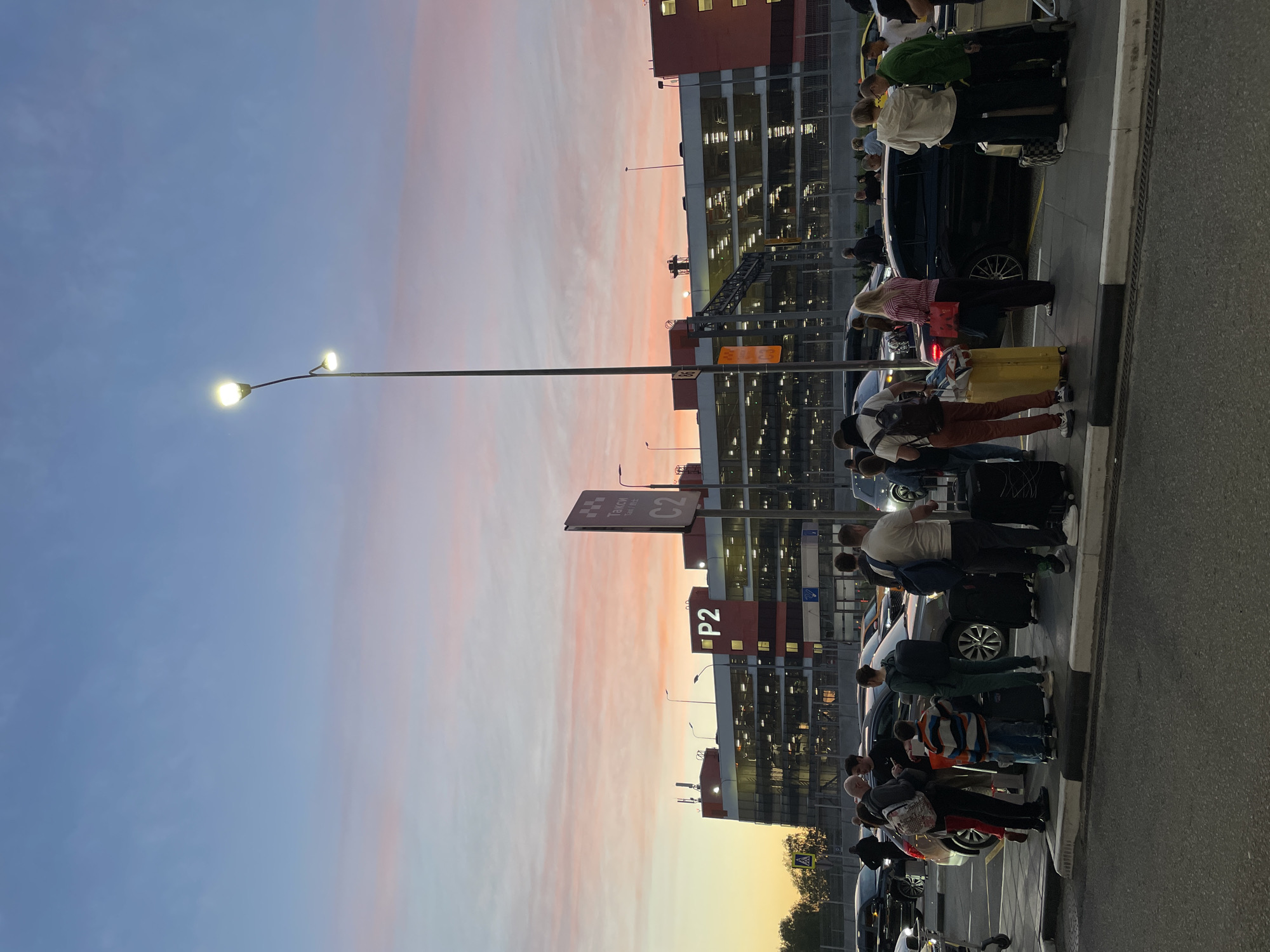 The image depicts a curbside scene at Sheremetyevo International Airport in Khimki, Russia. Passengers with luggage are standing on the sidewalk near a line of cars. A multi-level parking garage is visible in the background, marked with "P2" signage. Overhead, a taxi stand sign is identifiable with Cyrillic text. The sky shows a sunset gradient with pink and blue hues. A street lamp with two illuminated globes stands in the foreground.
