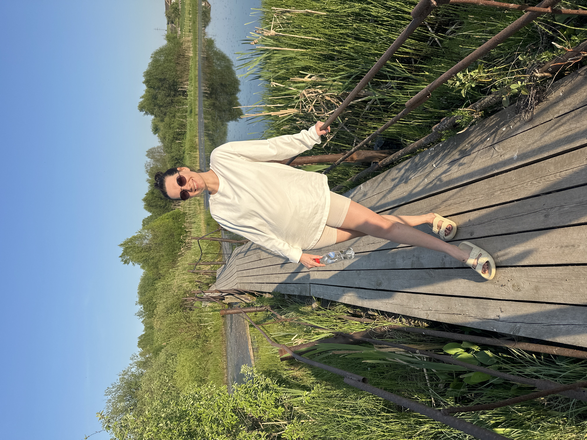 A woman in sunglasses, a white long-sleeved shirt, and beige shorts and sandals stands on a wooden boardwalk holding a plastic water bottle. She is grasping a rusty metal railing on the right side of the boardwalk. The boardwalk is elevated above a wetland area with tall grasses and a body of water visible in the background. The sky is clear and blue. Based on the given location, this scene could be in Альняш, Russia.