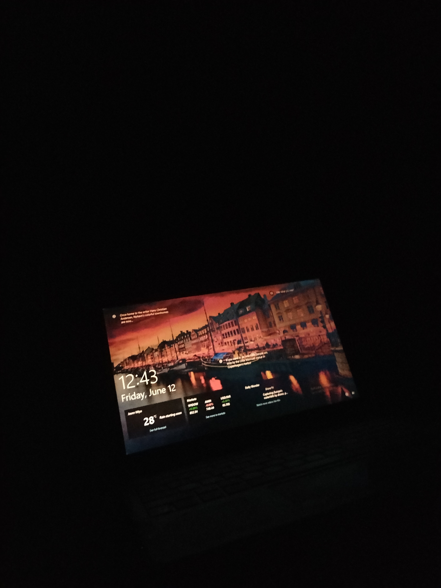 The image shows a laptop screen displaying a lock screen with a city view at sunset as the background. The time is 12:43 on Friday, June 12. There is also a weather widget showing 28°C and "Rain starting soon". Below this, there are some market updates and news headlines. The laptop is placed on a dark surface, likely a keyboard, and the surrounding environment is dark, suggesting the photo was taken in a dimly lit room or at night.