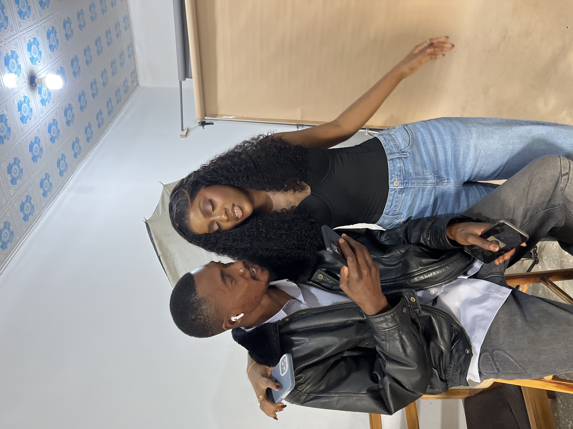 This image, set in an indoor location in the city of Gura Top, Nigeria, features two individuals: a man and a woman, standing closely together. The man, with a dark complexion, short dark hair, and a nose piercing on his left nostril, wears a black leather jacket with a dark faux fur collar over a light-colored collared shirt, and dark grey jeans. He has a white wireless earbud in his right ear and holds a black smartphone in his left hand, looking down at its screen. The woman, also with a dark complexion, wears a long, dark, curly wig, a black fitted top, and light blue wide-leg jeans. Her left arm is draped over the man's left shoulder, and she holds another black smartphone, resting it on his shoulder. She is also looking down, presumably at the man's phone, with her lips slightly parted. Her right arm hangs loosely at her side.
The background elements include a beige seamless paper backdrop rolled onto a stand on the right, and a white wall on the left with part of a photography softbox or umbrella visible. The ceiling is white, adorned with blue floral or scrollwork patterned tiles, from which a single illuminated bare light bulb hangs. The floor appears to be a light-colored concrete or industrial surface. A wooden stool or chair is partially visible beneath the man. The lighting is bright and direct, typical of an indoor studio setting.