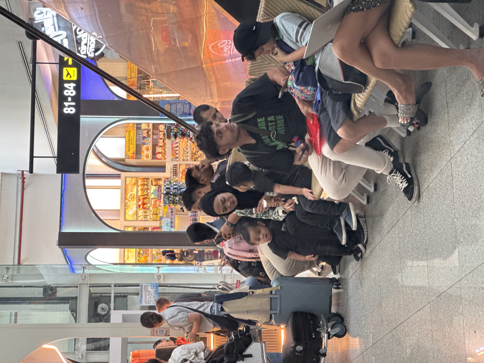 The image captures a scene inside an airport terminal in Mai Khao, Thailand. In the foreground, a group of people are seated on benches, waiting. A young child in a black t-shirt is looking towards the camera. To the right, a boy in a light blue shirt and a black cap is focused on a laptop, while an adult in a black t-shirt with green text is holding a phone. To the left, a woman in a patterned hijab is holding a phone, and a younger child in a black t-shirt is sitting beside her. Luggage is visible on the floor, including a dark suitcase and a light blue trolley bag. In the background, a shop with various packaged food items is displayed behind arched openings, and a digital sign shows "81-84" with a plane icon, indicating gate information. Escalators and glass railings are also part of the architectural environment. The lighting suggests an indoor setting with artificial illumination.