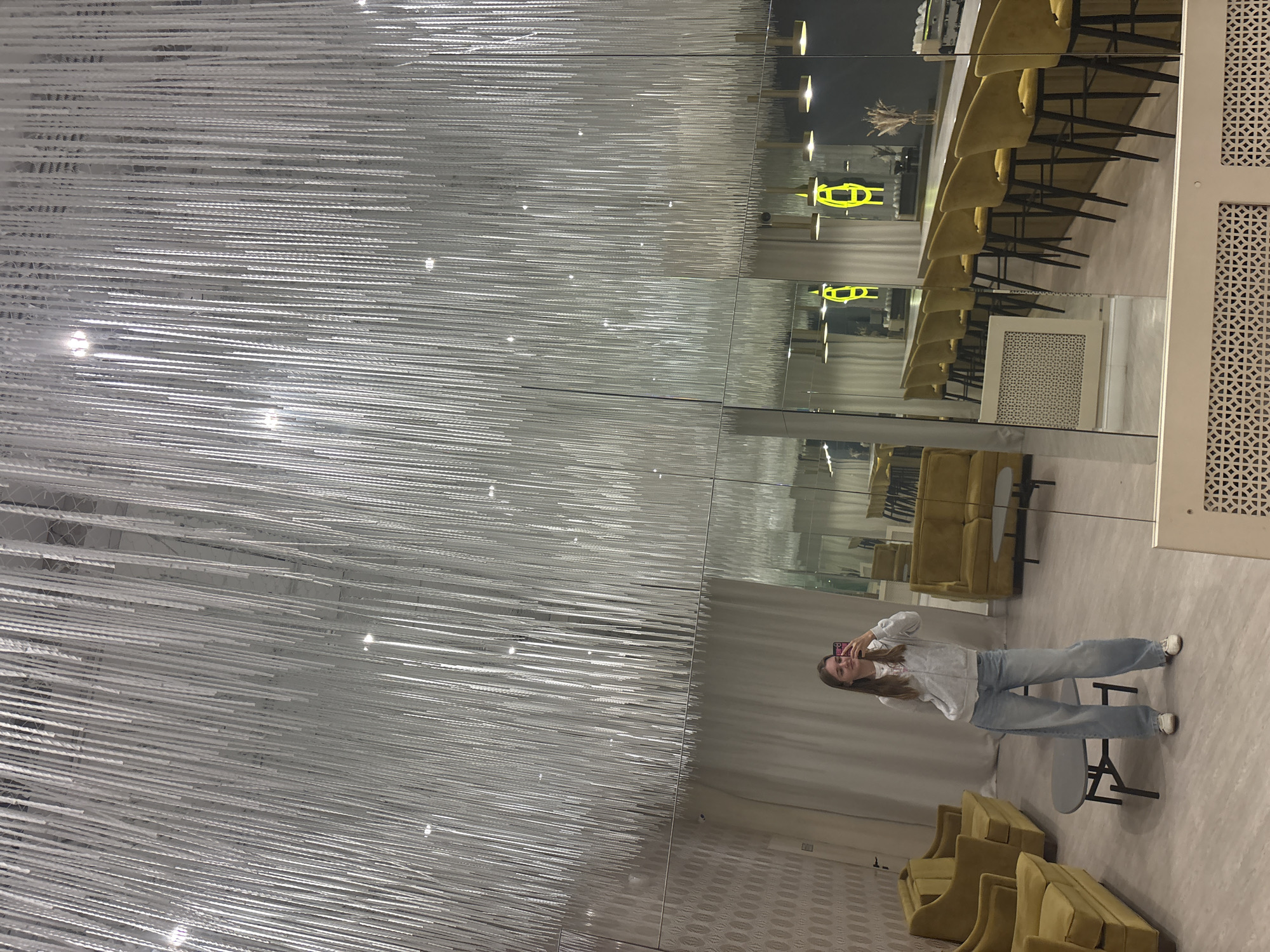 The image captures a woman reflected in a mirrored wall. The woman is holding her phone up as if taking a photo, and is wearing a white sweater and light blue jeans. The mirrored wall shows a room with yellow seating, a long table with high-backed chairs, and decorative lighting. A striking chandelier composed of multiple white strands hangs from the ceiling. The scene appears to be located in a restaurant or lobby. Based on other available context, the location is likely in Perm, Russia.