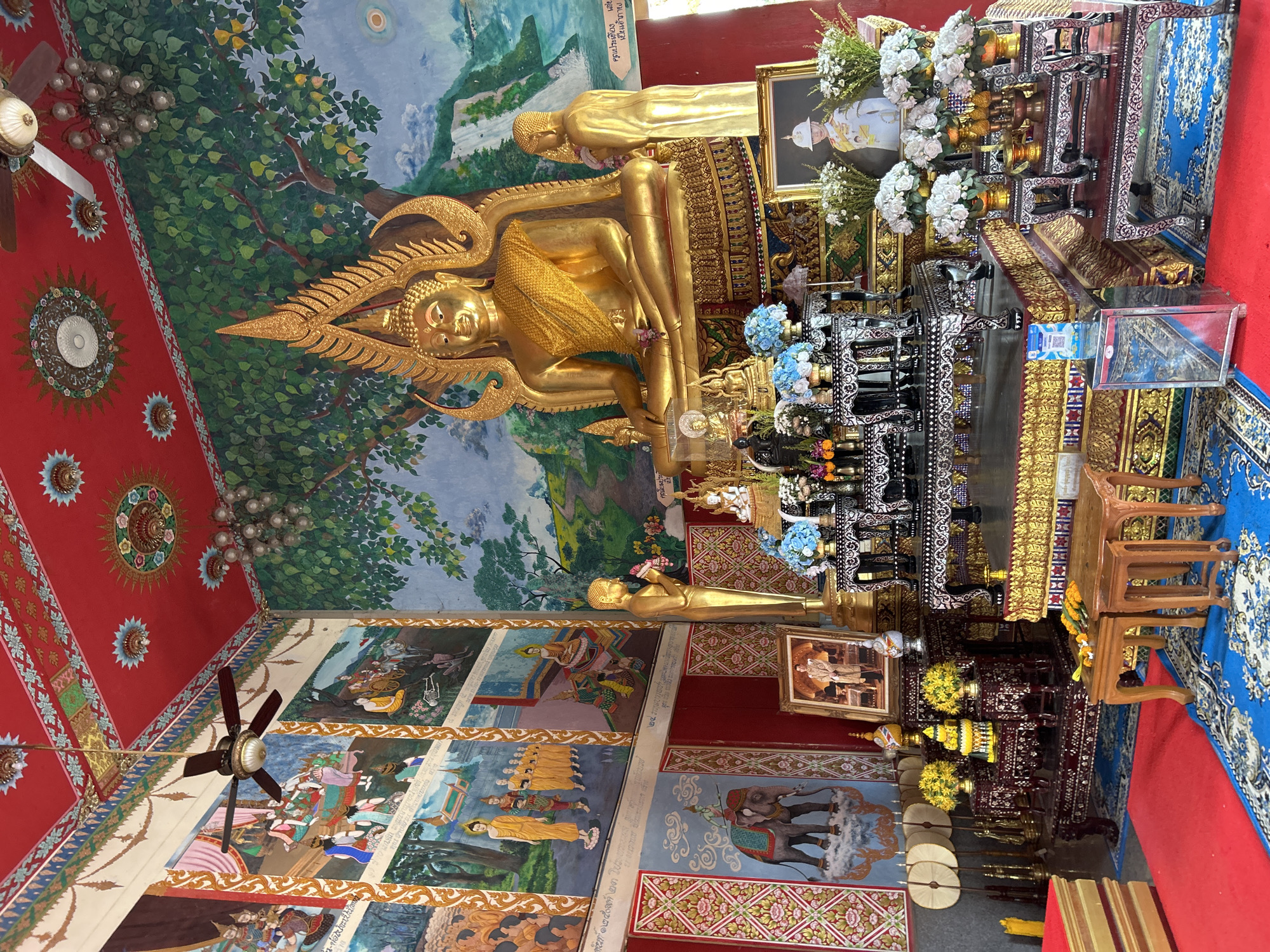 This image captures the interior of a Buddhist temple in Ko Samui District, Thailand. A large, gilded Buddha statue sits prominently on a tiered altar adorned with flowers and offerings. To the right of the main statue, a smaller gilded statue of a monk stands in prayer. The walls of the temple are decorated with vibrant murals depicting Buddhist stories and scenes, including elephants and processions. The ceiling is painted red and features intricate patterns, with a ceiling fan and a chandelier visible. The floor is covered with a red carpet and a blue rug with a floral pattern. In the foreground, there are wooden stools and a small shrine with more flowers and offerings. A framed portrait of a man in ceremonial attire is placed on the altar. The overall atmosphere suggests a place of worship and cultural significance.