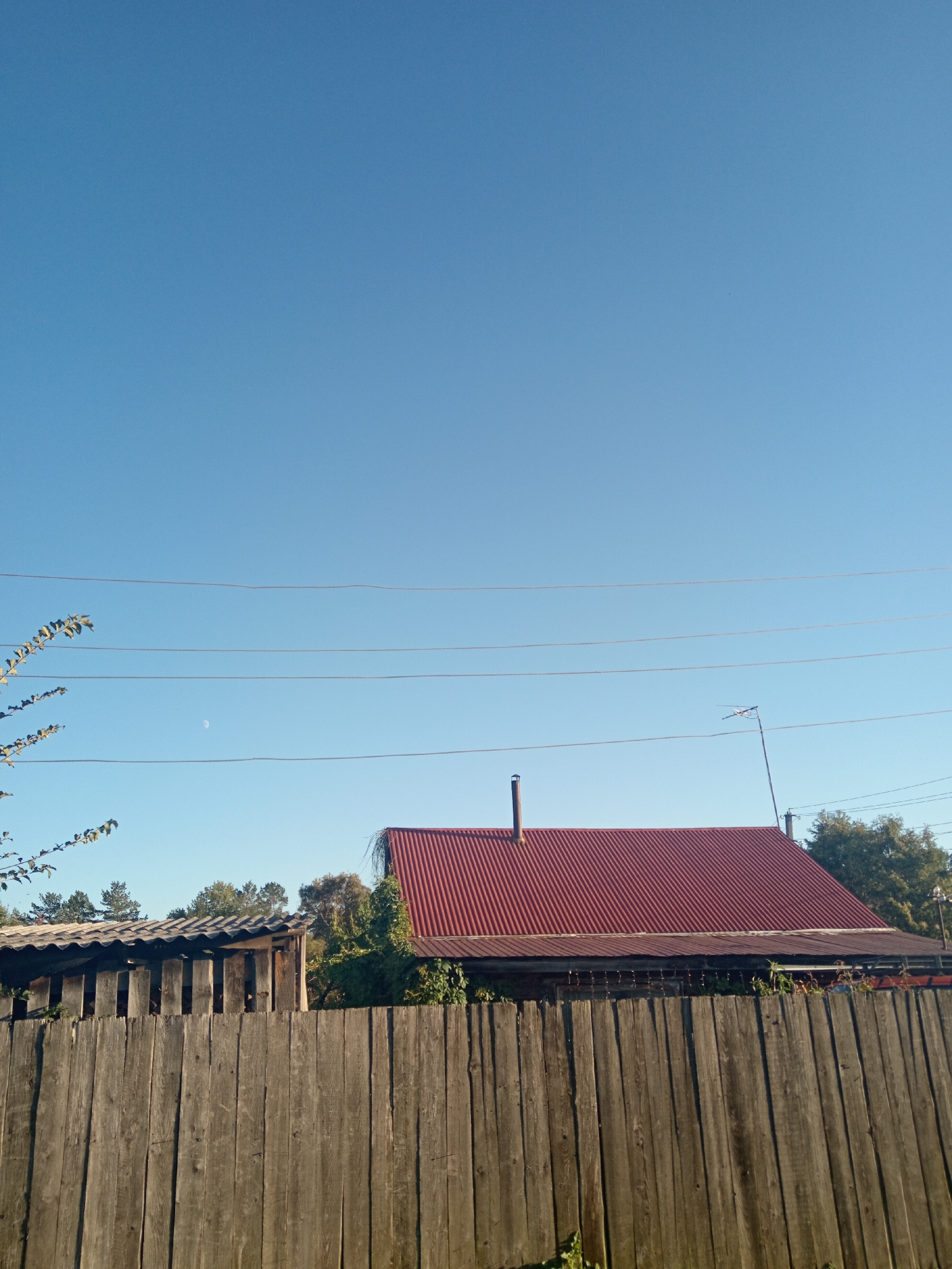 A high-angle, full outdoor shot shows a rustic scene under a clear blue sky, likely in Novopokrovka, Russia. In the foreground, a weathered wooden fence stretches horizontally across the frame, its vertical planks showing signs of age. Behind the fence, a corrugated red roof of a building is visible, with a chimney. To the left of this structure, another smaller building with a wavy, gray roof can be seen. Trees and greenery are scattered throughout, with some foliage encroaching on the buildings. Several power lines are strung across the sky, and a small, crescent moon is faintly visible. The overall impression is one of a rural or semi-rural setting with simple, aged structures and natural elements.